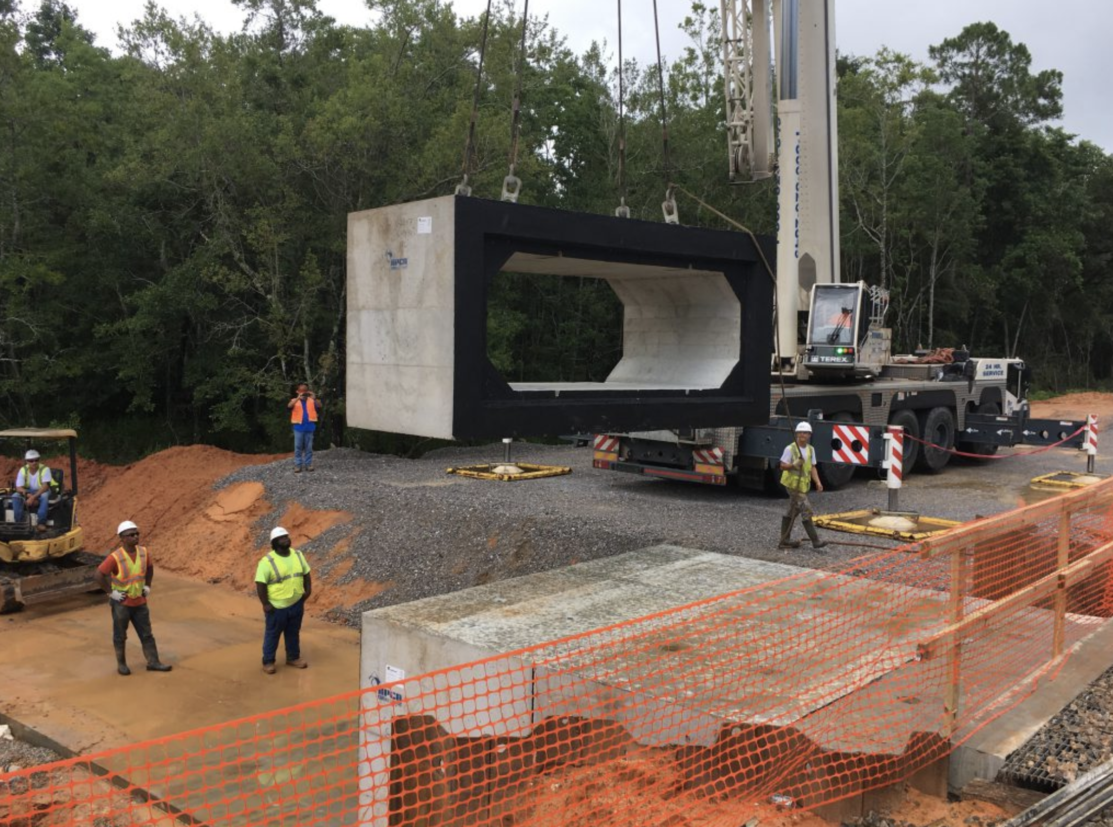 Construction workers and equipment installing a large gray concrete tunnel segment on a construction site with trees in the background.