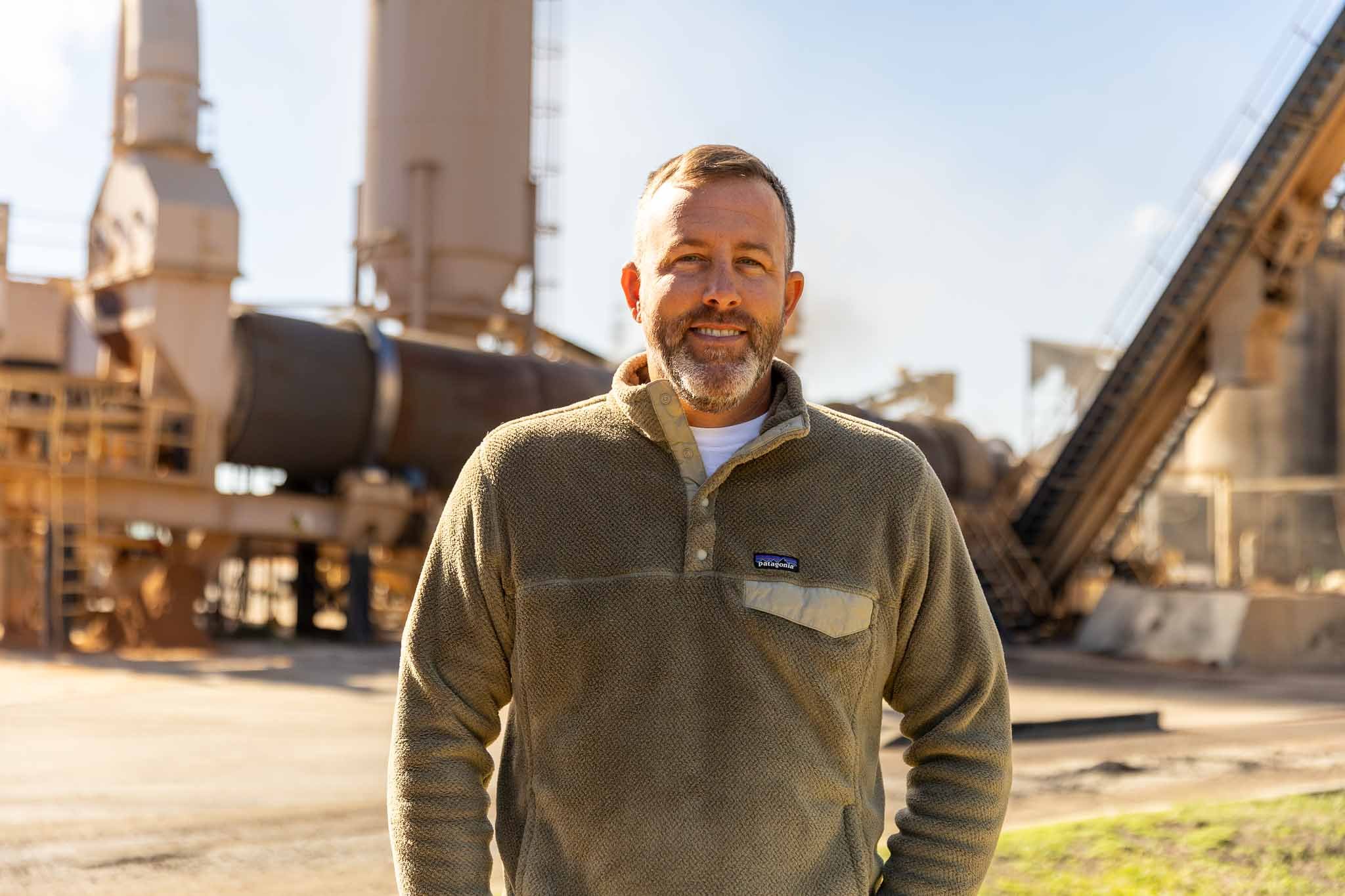 A man standing outdoors in front of industrial equipment, smiling at the camera. He is wearing a brown fleece pullover and has short, gray hair and a beard.