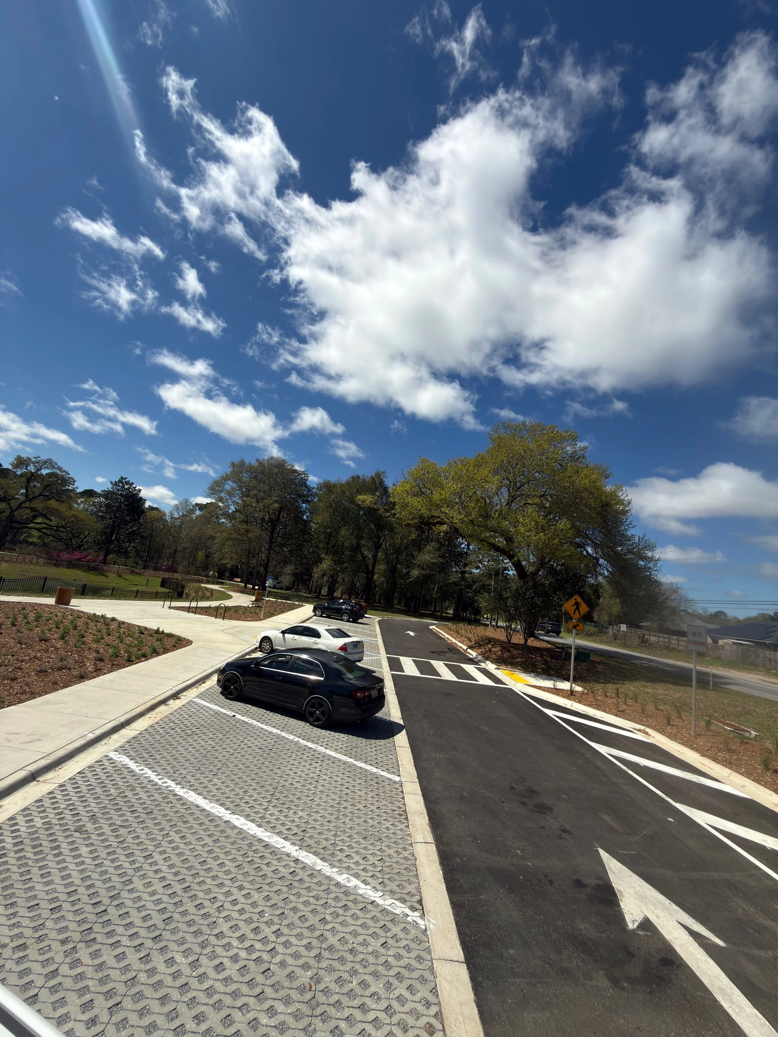 A parking lot with some parked cars, a sidewalk, trees, a cloudy sky, and a pedestrian crossing sign.