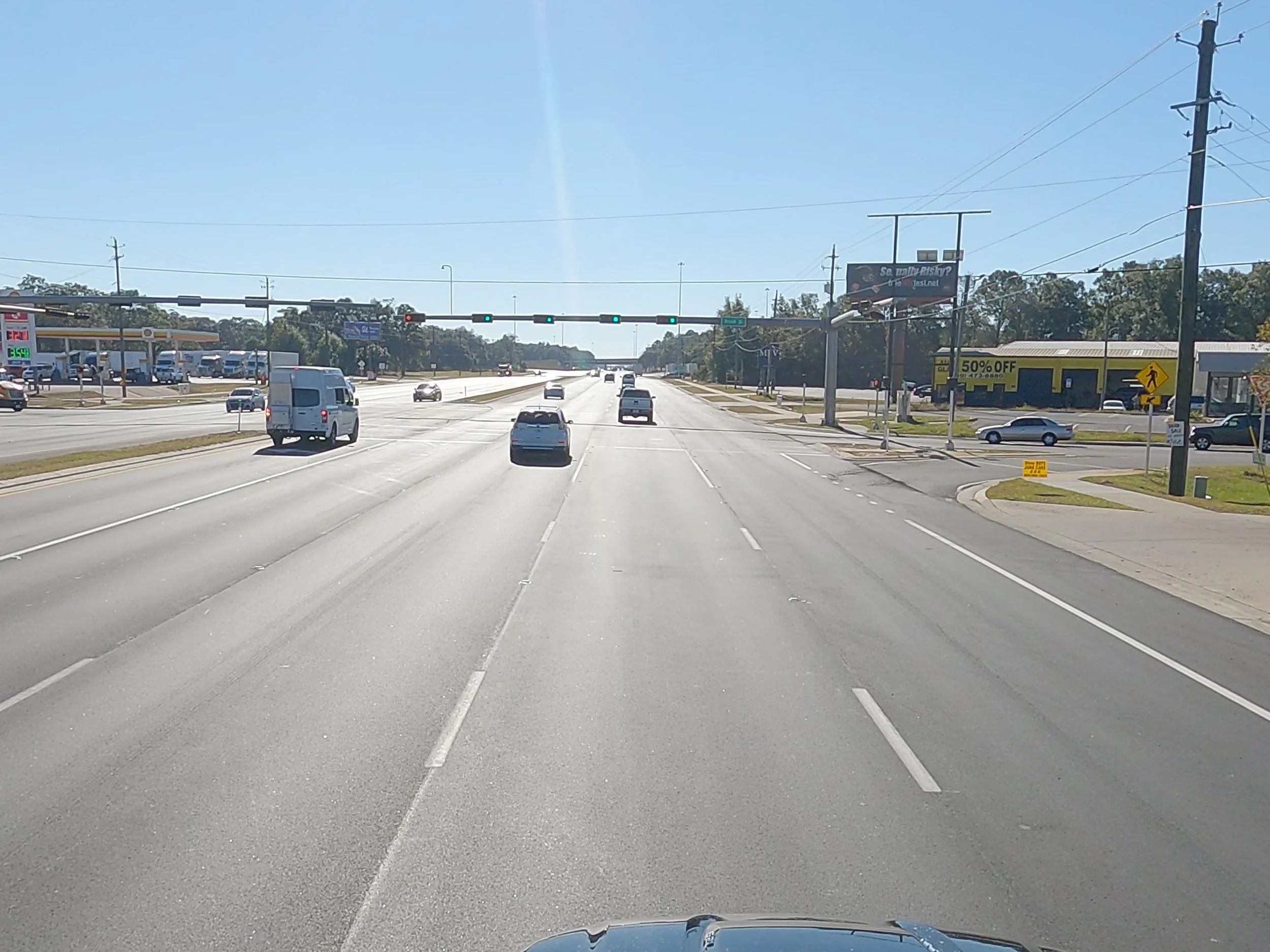 A multi-lane intersection with a green traffic light, a gas station on the left, and a shopping center with a yellow sign on the right. The road has light traffic and clear blue skies.
