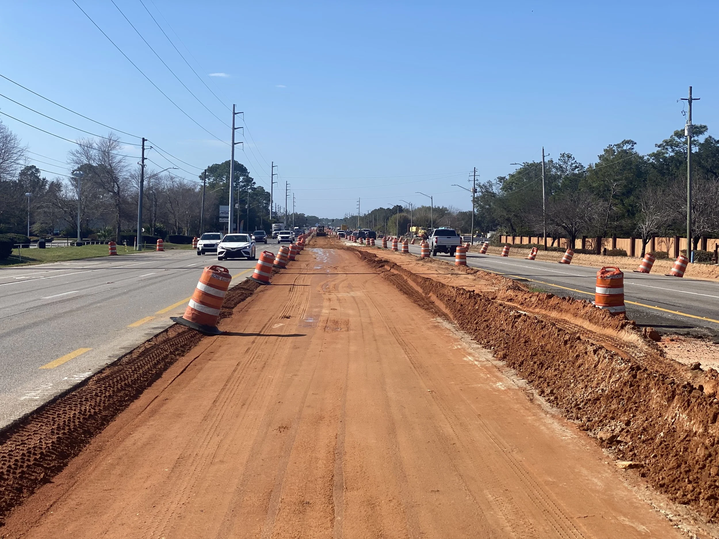 Road construction with orange traffic barrels and dirt path in the middle, cars driving on the left and right lanes, trees and utility poles along the road under a clear blue sky.