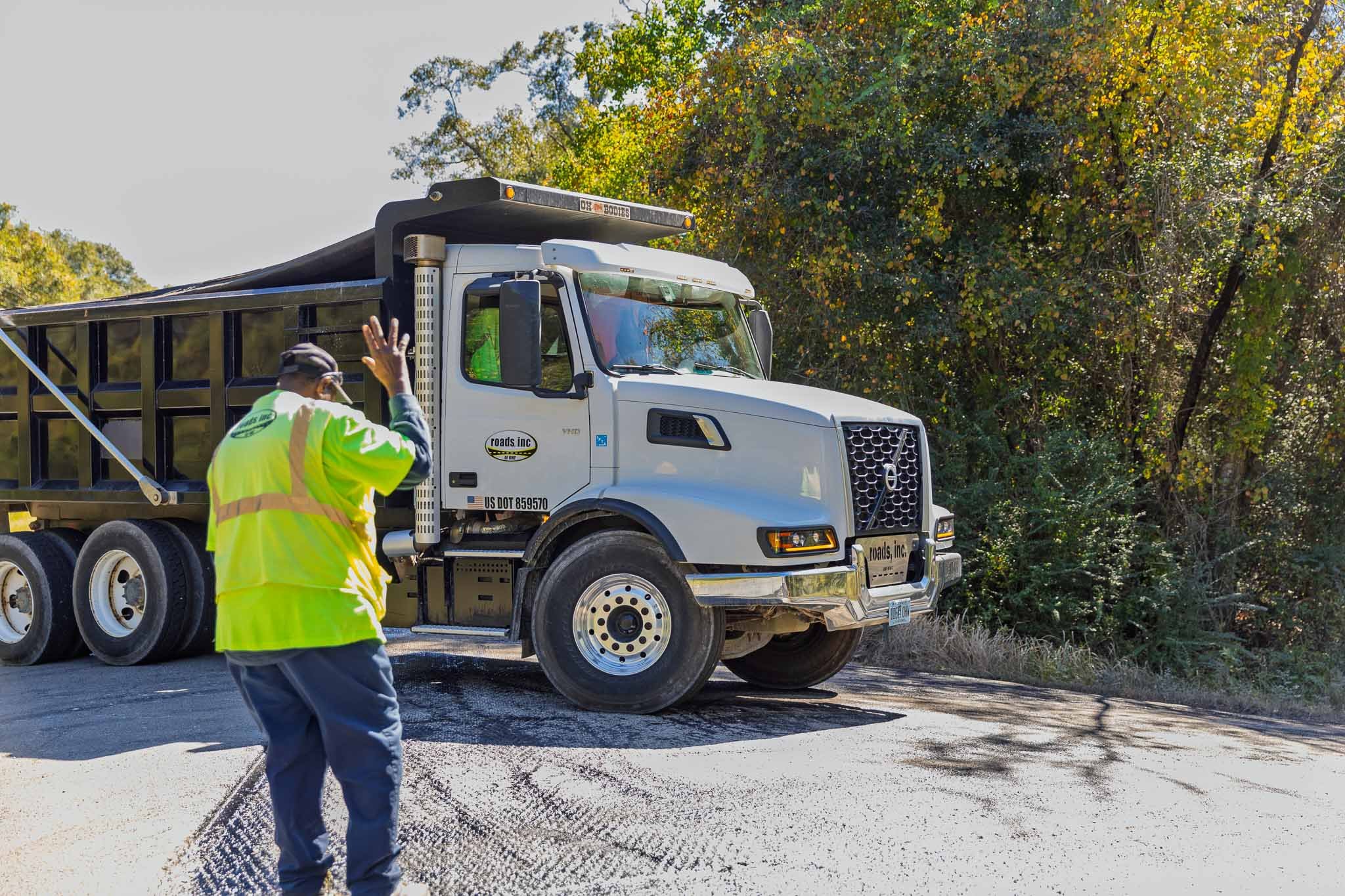A worker wearing a neon yellow safety vest and dark blue pants signals a large dump truck on a road, with trees displaying fall foliage in the background.