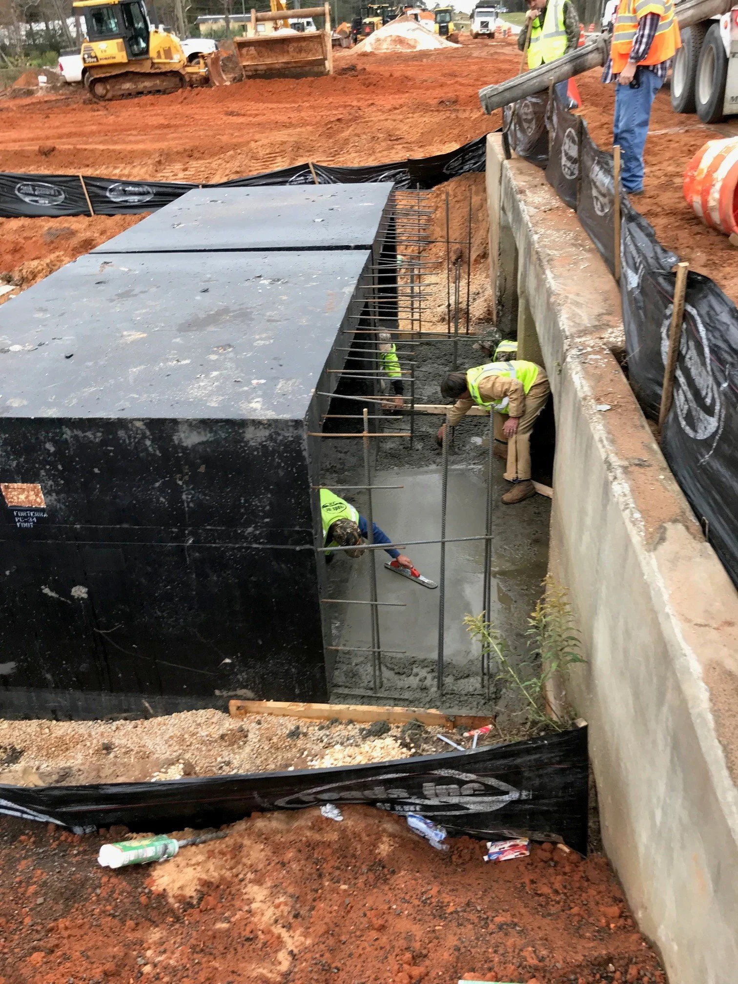 Construction workers are pouring concrete in a trench at a construction site. There is heavy machinery and orange dirt in the background, and black barriers and a concrete wall surround the work area.