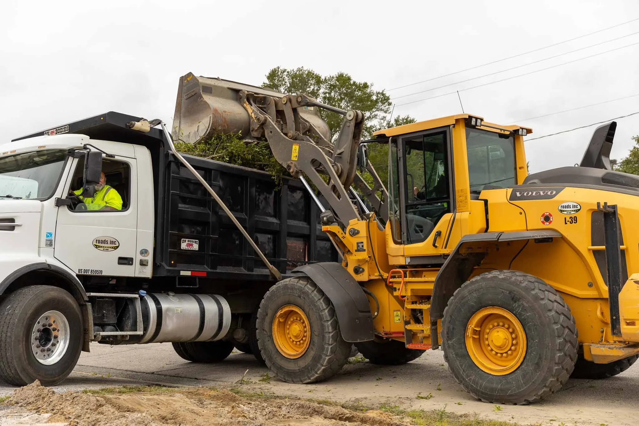 Construction vehicles on a road, including a large yellow Volvo loader and a white dump truck, with a worker visible inside the truck's cab.