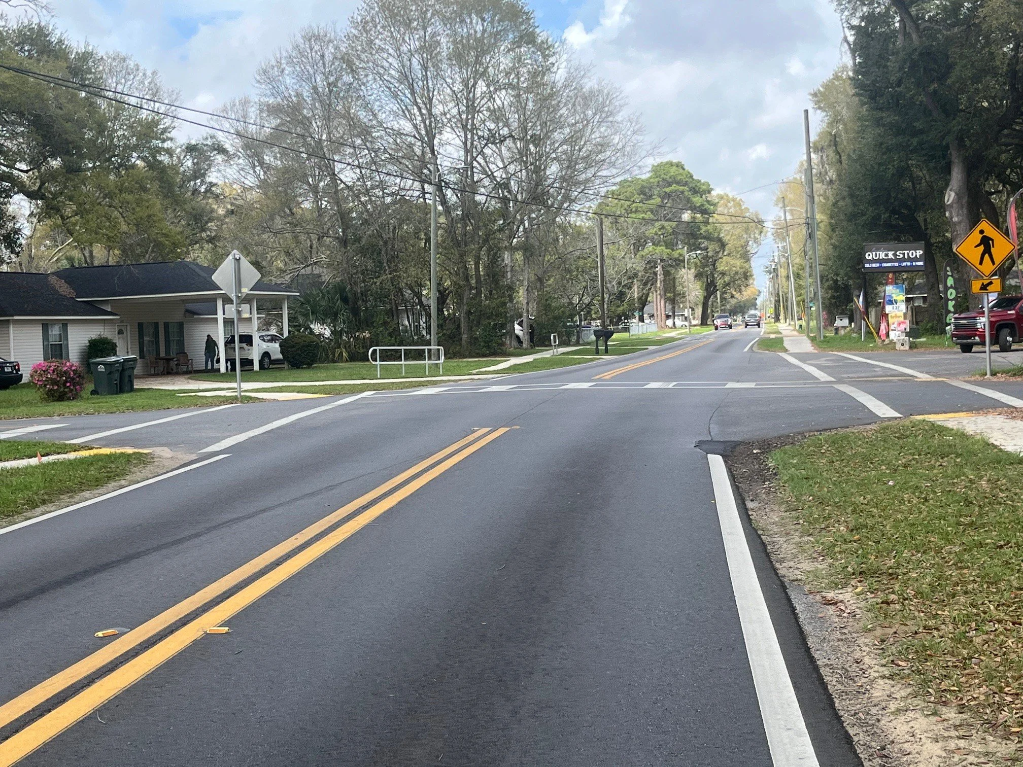 A two-lane street in a suburban area with trees, houses, and a crosswalk. A pedestrian sign and a quick stop sign are visible on the right side of the street.