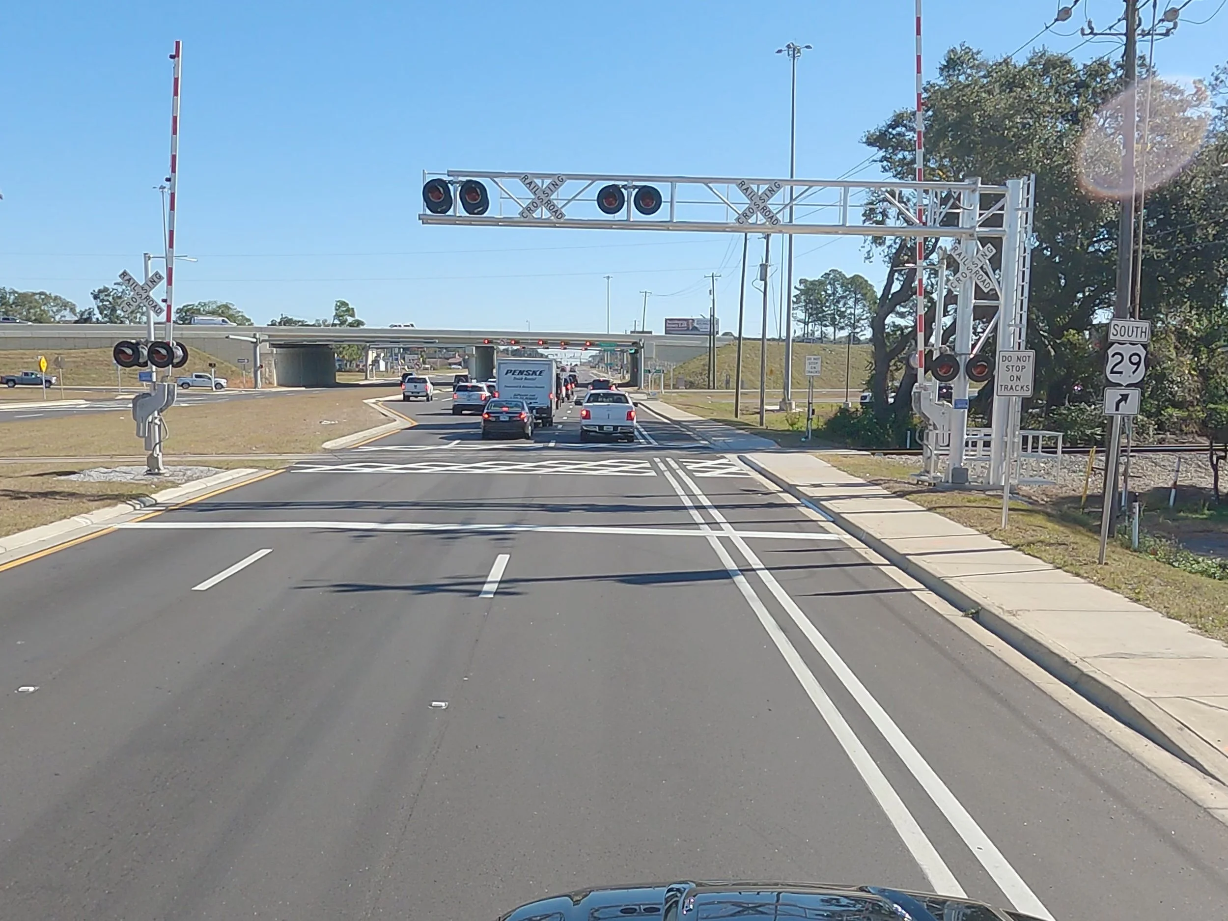 A roadway with multiple lanes approaching a crossing gate for a railroad. Cars are stopped at the crossing, and there is a bridge overpass in the background. Emergency lights are mounted on the crossing gate structure.