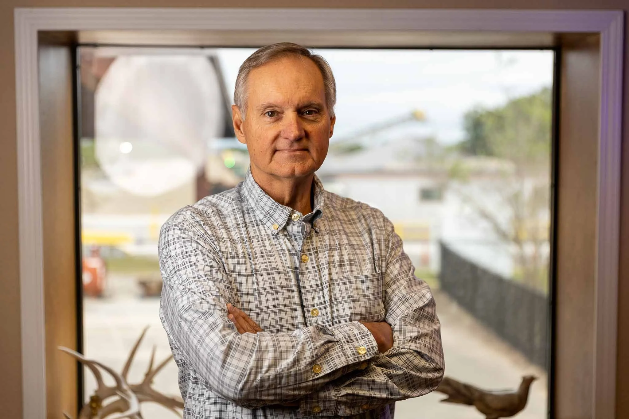 An older man with crossed arms standing in front of a window, wearing a checkered shirt, with an outdoor scene in the background.