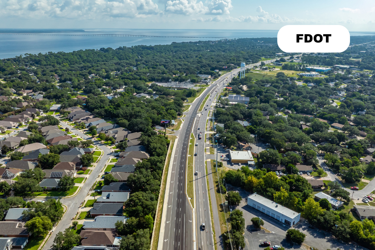 An aerial view of a suburban area with a highway running through it, surrounded by residential houses, trees, and some commercial buildings, near a body of water in the background. The image has a label saying "FDOT".