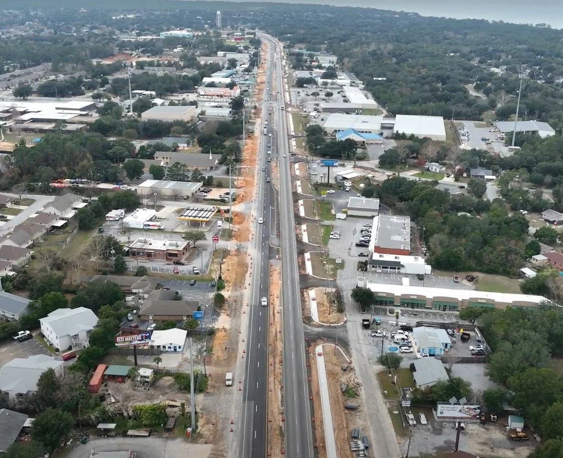 An aerial view of a highway under construction running through a suburban area with houses, commercial buildings, and trees on either side.