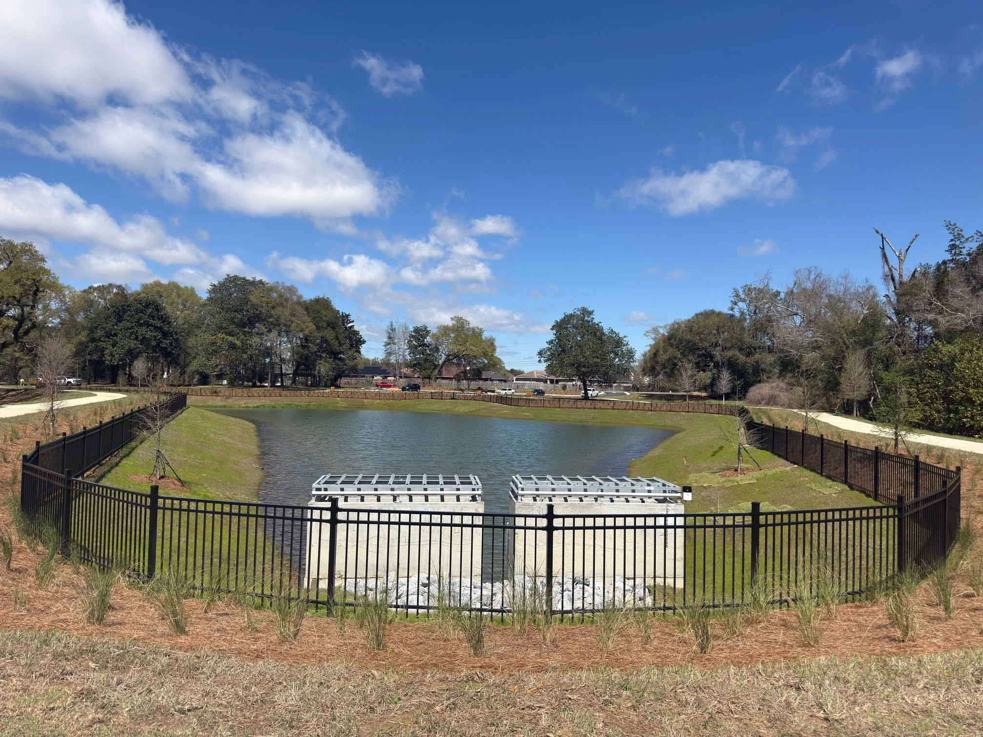 A small pond surrounded by a black fence with two concrete water spillways in the front. The sky is blue with scattered clouds, and trees line the background with a few cars visible beyond the trees.