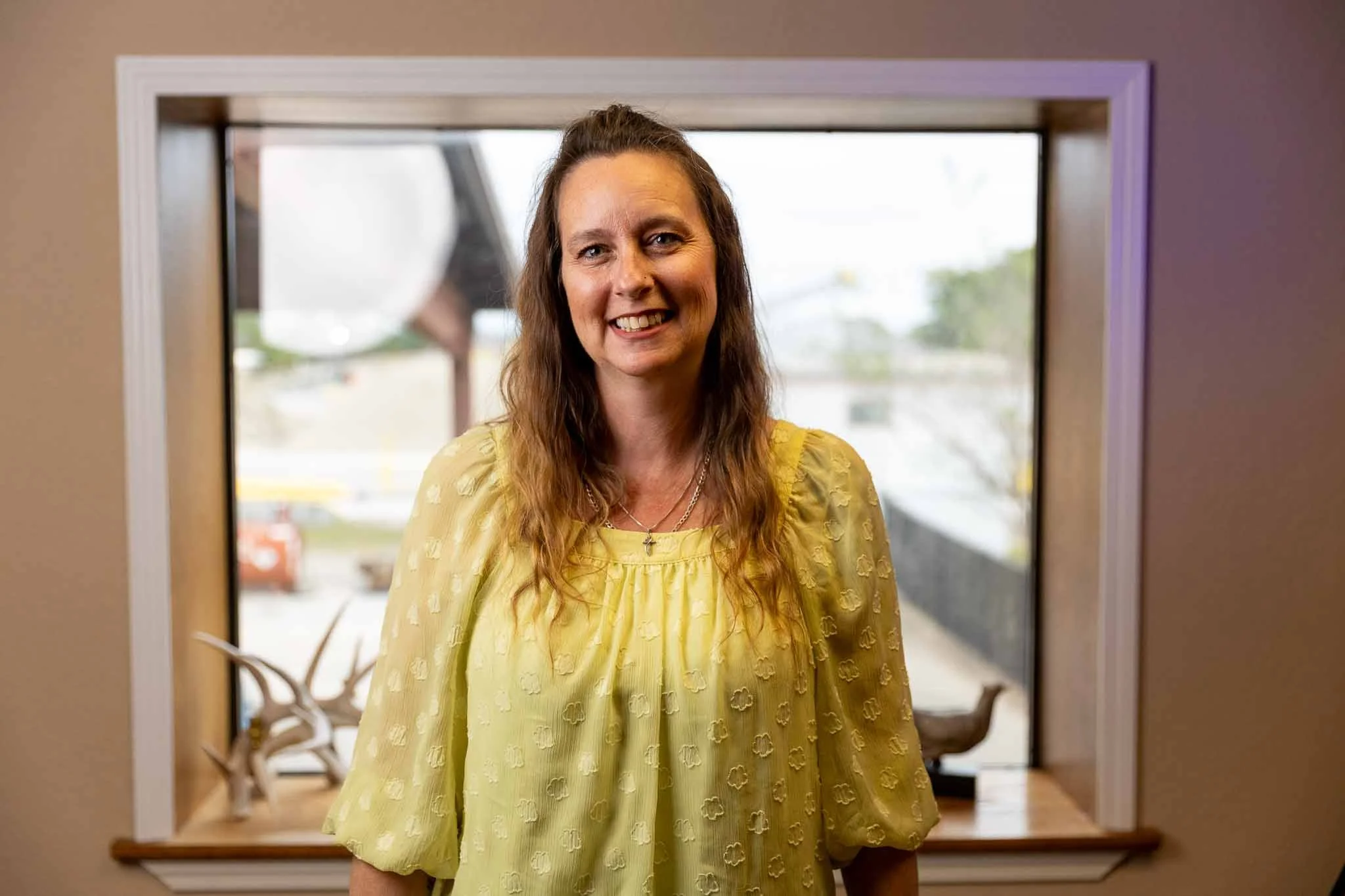 A woman with long curly brown hair smiling, wearing a light yellow blouse with embroidered flower patterns, standing in front of a window with a view of a parking lot and some trees outside.