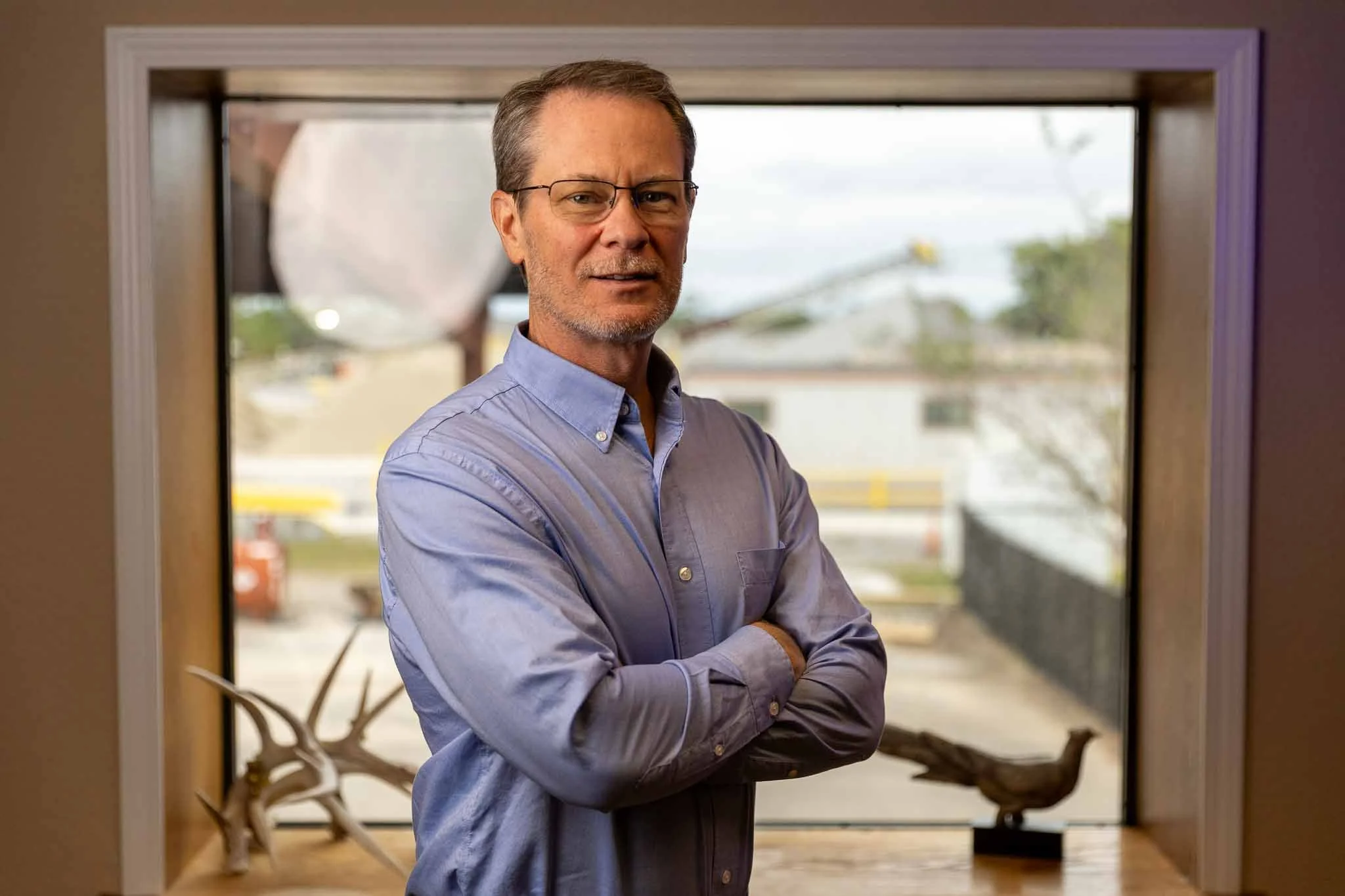 A middle-aged man wearing glasses and a light blue button-up shirt stands with his arms crossed in front of a window, with a wooden sculpture and antlers on a table behind him.