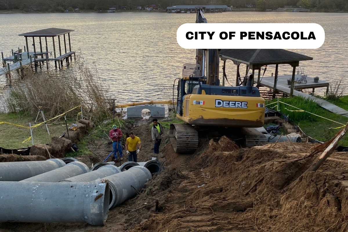 Construction workers installing large drainage pipes near the water's edge by a lake in Pensacola. An excavator labeled 'Deere' is present, and a gazebo and dock are visible on the lakeshore.