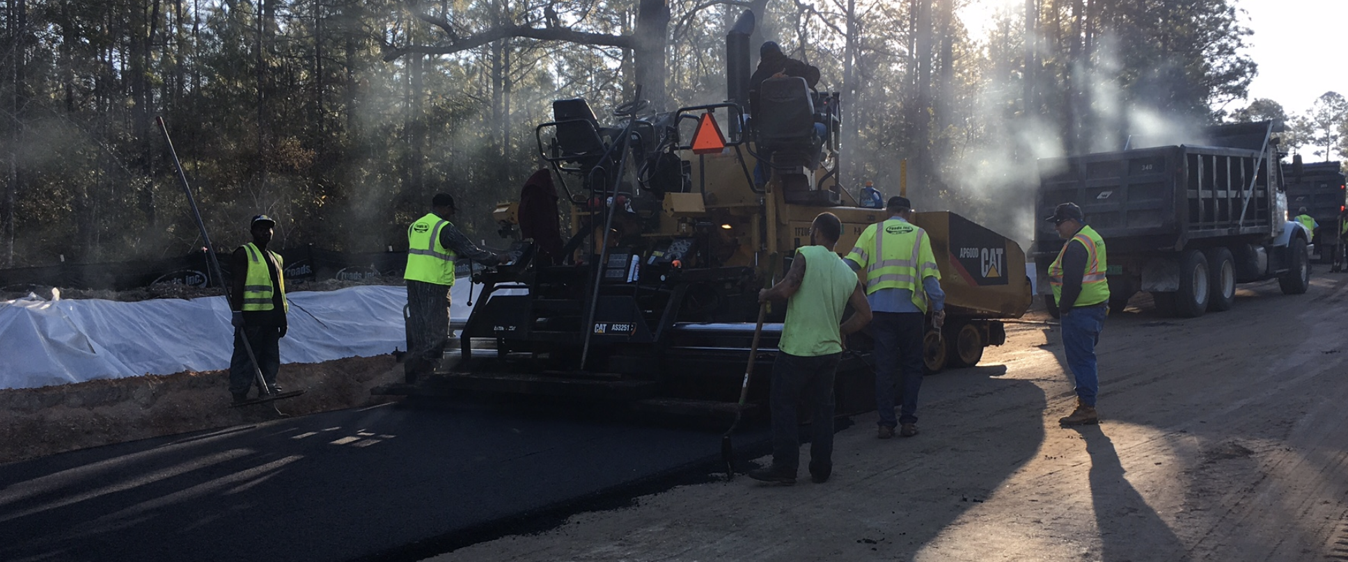Construction workers in safety vests paving a road with asphalt, surrounded by trucks and machinery, in a wooded area during daylight.