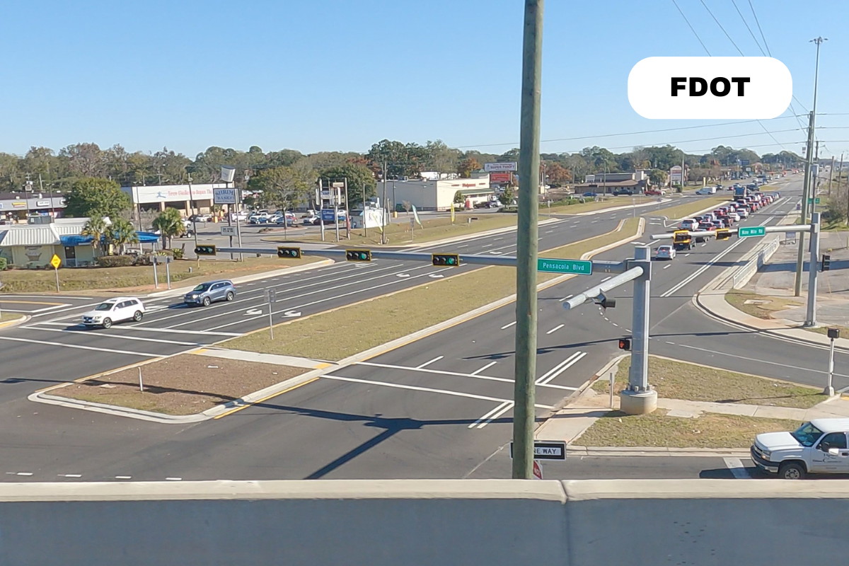 A busy intersection with multiple lanes of traffic, traffic lights, and street signs for Pensacola Blvd and nearby roads. There are cars driving in different directions and a small grassy median. The sky is clear and blue.