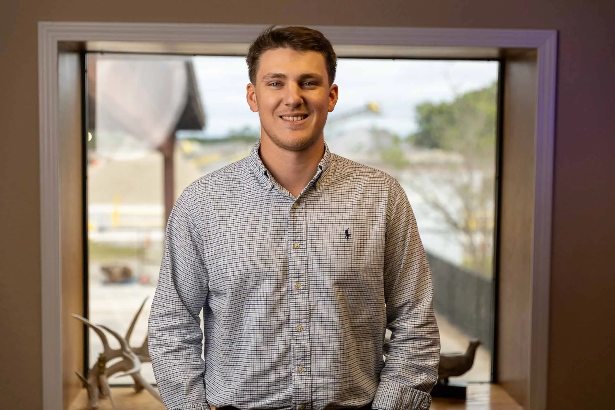 A young man with short brown hair, wearing a light-colored checkered shirt, smiling and standing indoors in front of a window with a blurred outdoor scene.