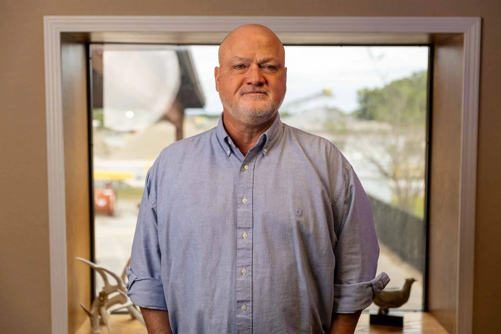 A middle-aged man with a bald head and beard, wearing a light blue button-up shirt, standing inside near a window with an outdoor parking lot visible in the background.