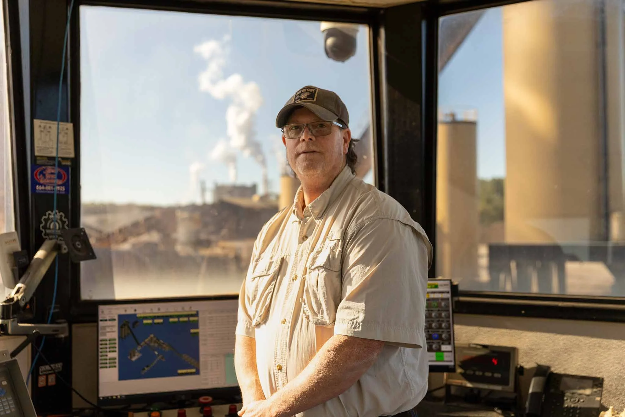 A man wearing glasses, a beige shirt, and a baseball cap standing inside a control room with industrial equipment and monitors, with a large window showing a power plant and smokestacks in the background.