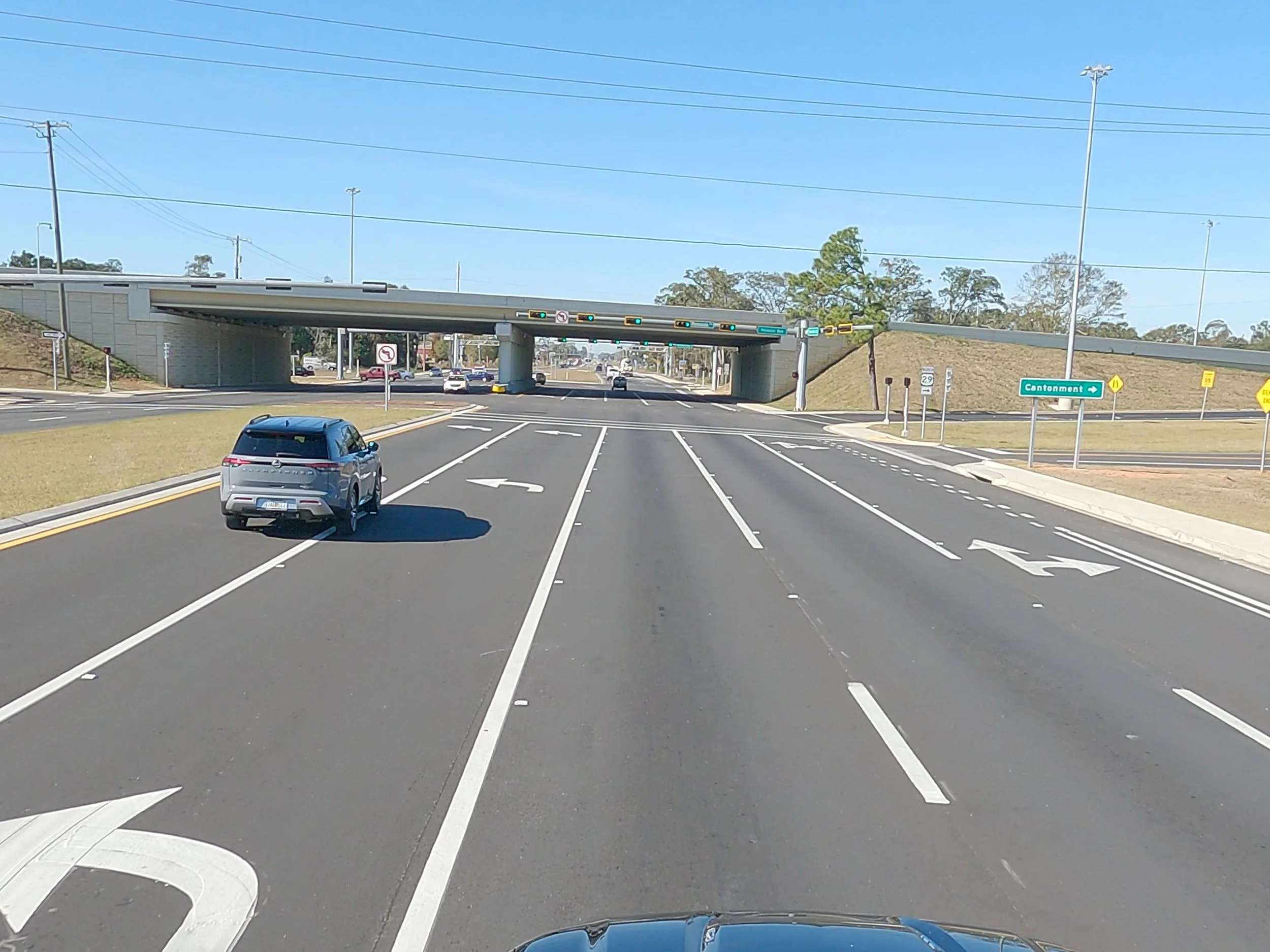 Empty multi-lane road with a car in the left lane approaching an overpass, with directional signs indicating CANTONMENT and other traffic signs, under a clear blue sky.