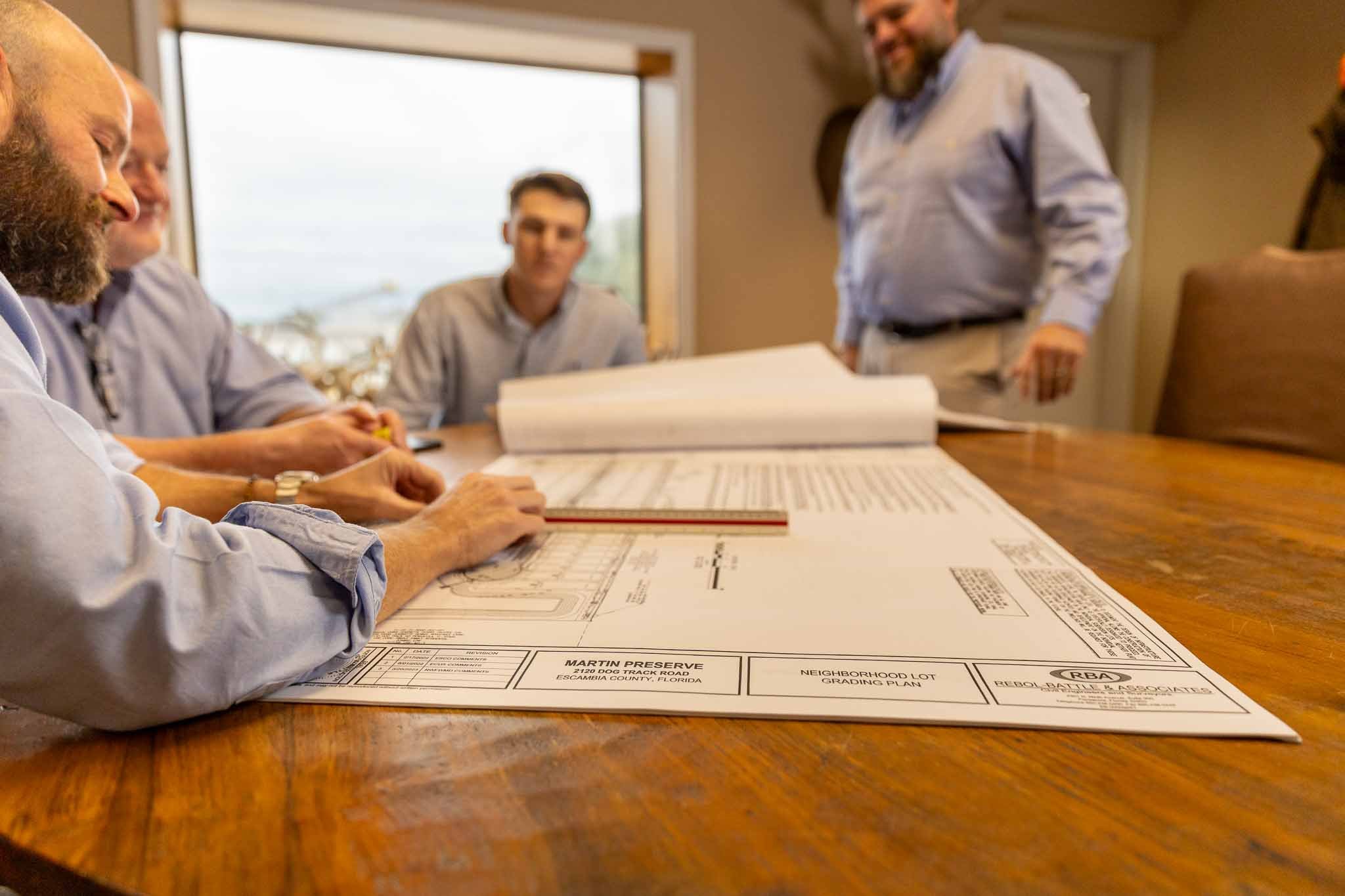 A group of four men in a meeting room discussing architectural blueprints on a wooden table, with large windows in the background.