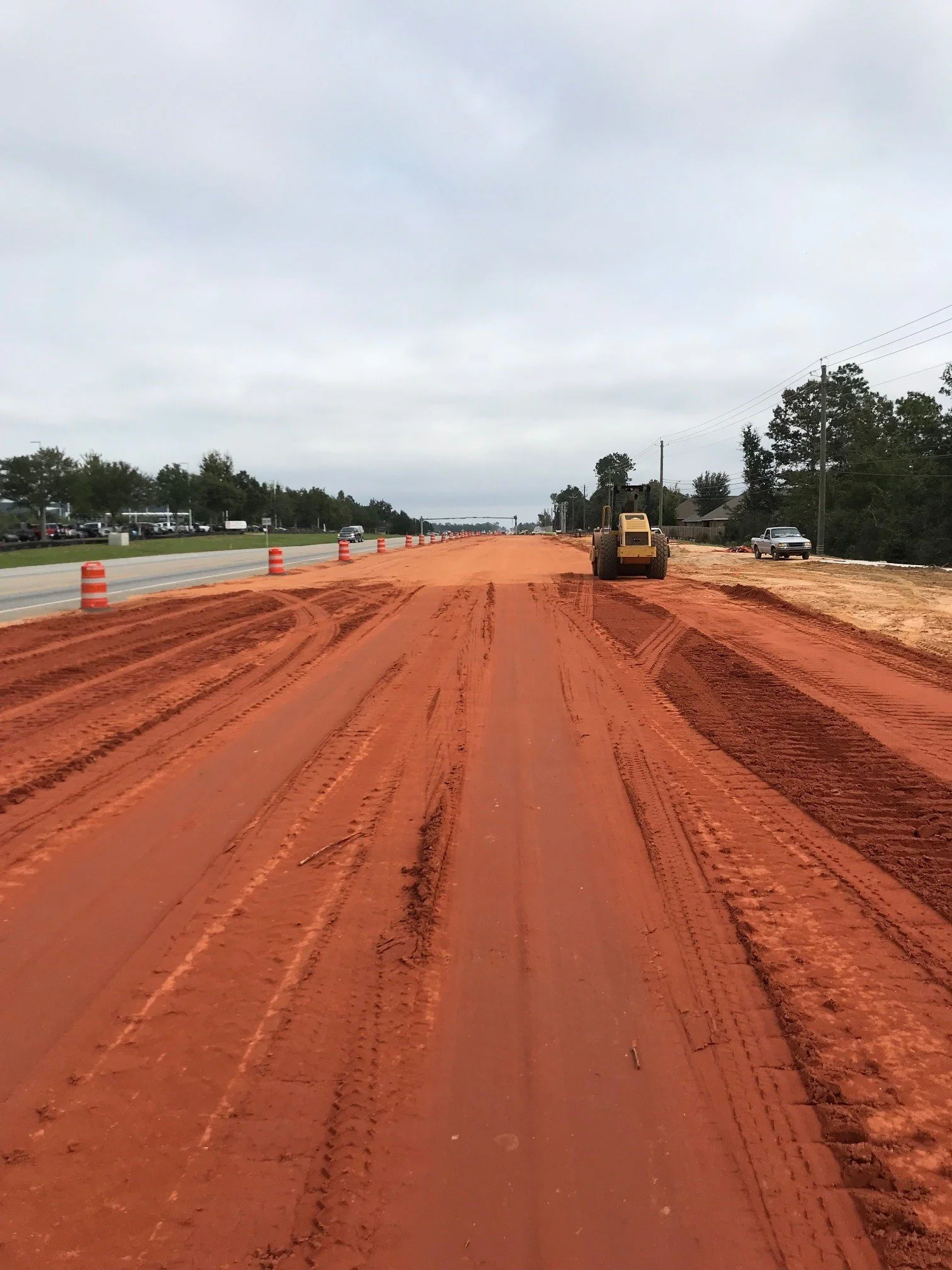 Construction site with a bulldozer on a dirt road, orange safety cones along the side, and vehicles on the paved road to the left.