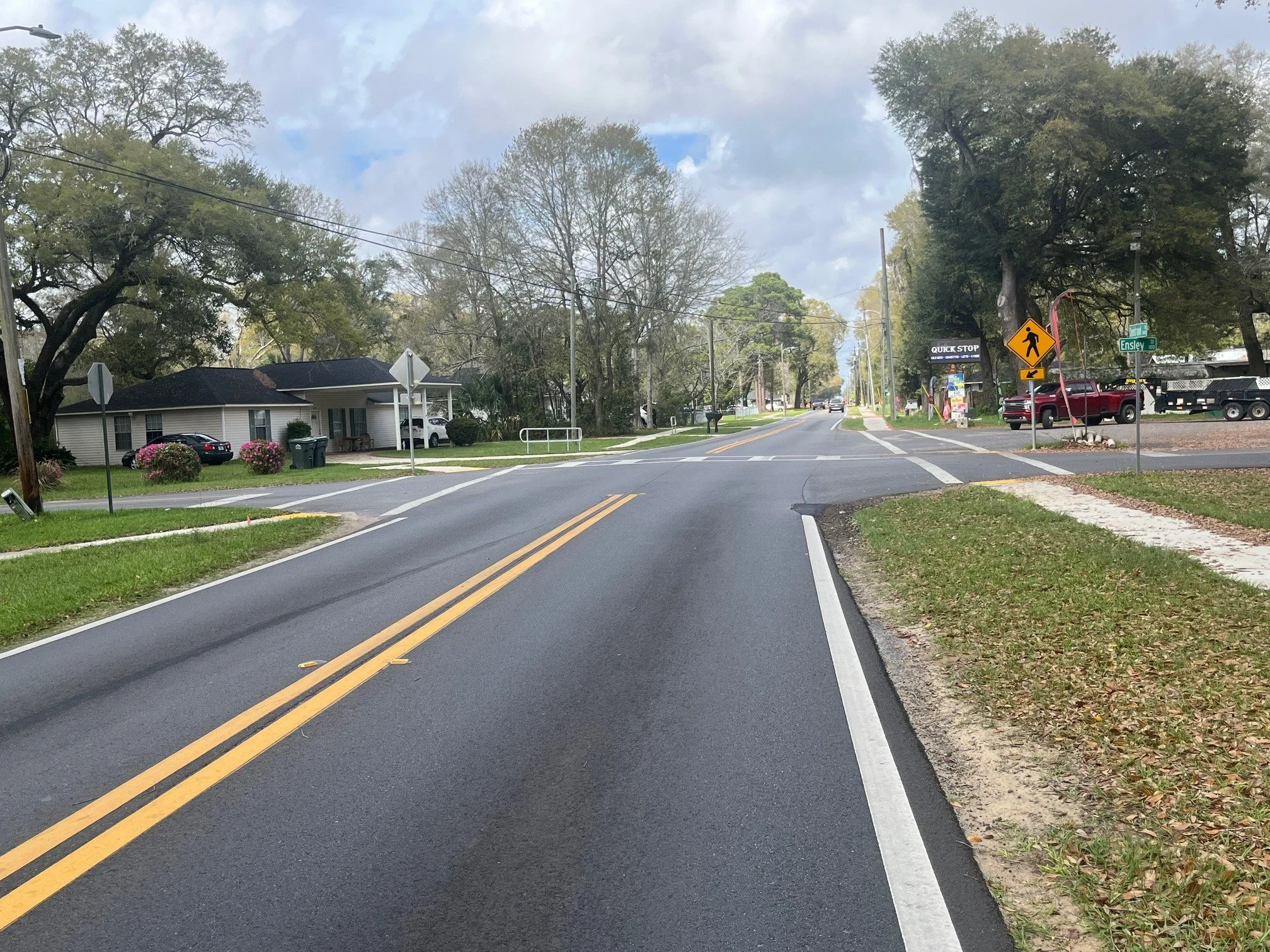 A suburban street with trees, residential houses, and cars parked along the side. There are traffic signs, a crosswalk, and a sidewalk on the right side of the street.