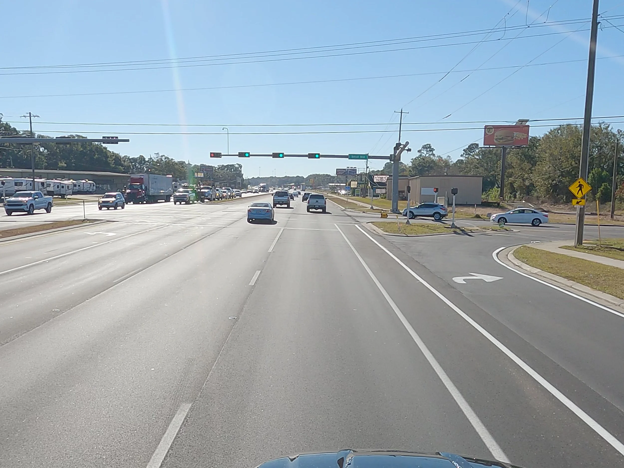 A busy multi-lane street with cars, trucks, and SUVs traveling in both directions. Traffic lights are green, and there are commercial buildings, billboards, and a walking pedestrian sign on the right side.