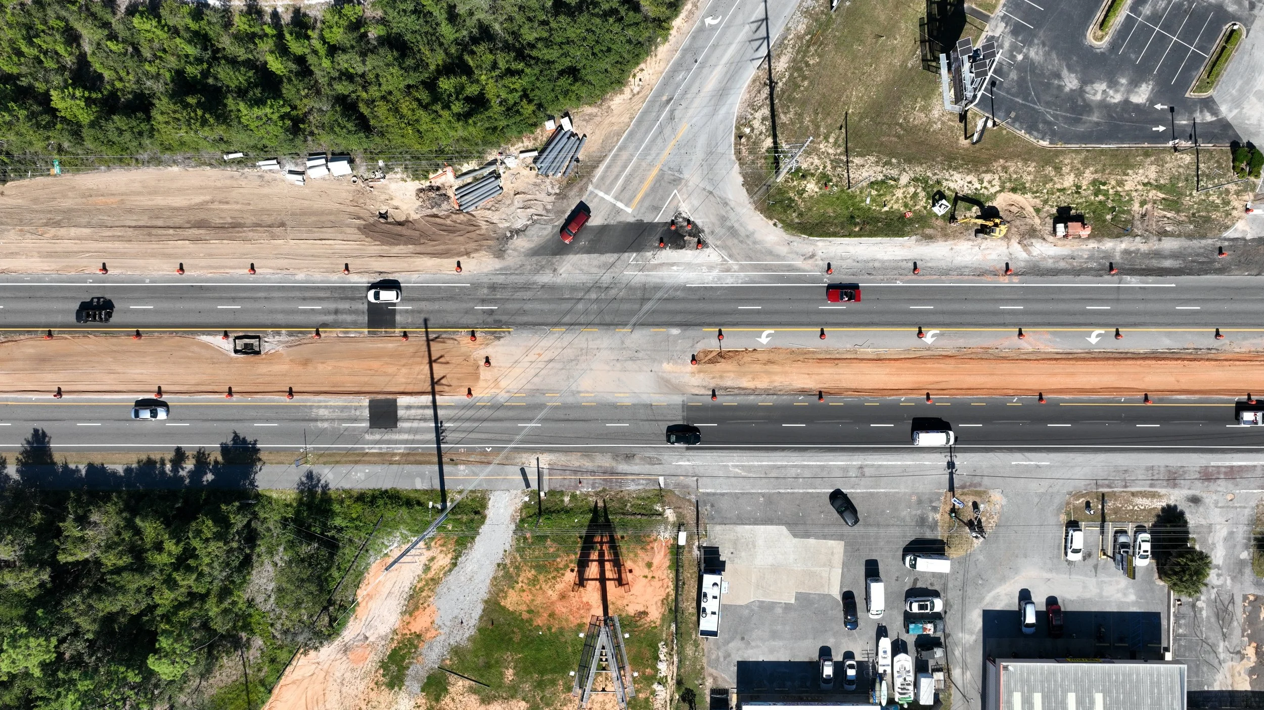 An aerial view of a multi-lane road with construction work in progress, traffic cones, and vehicles, along with parking lots and buildings nearby.