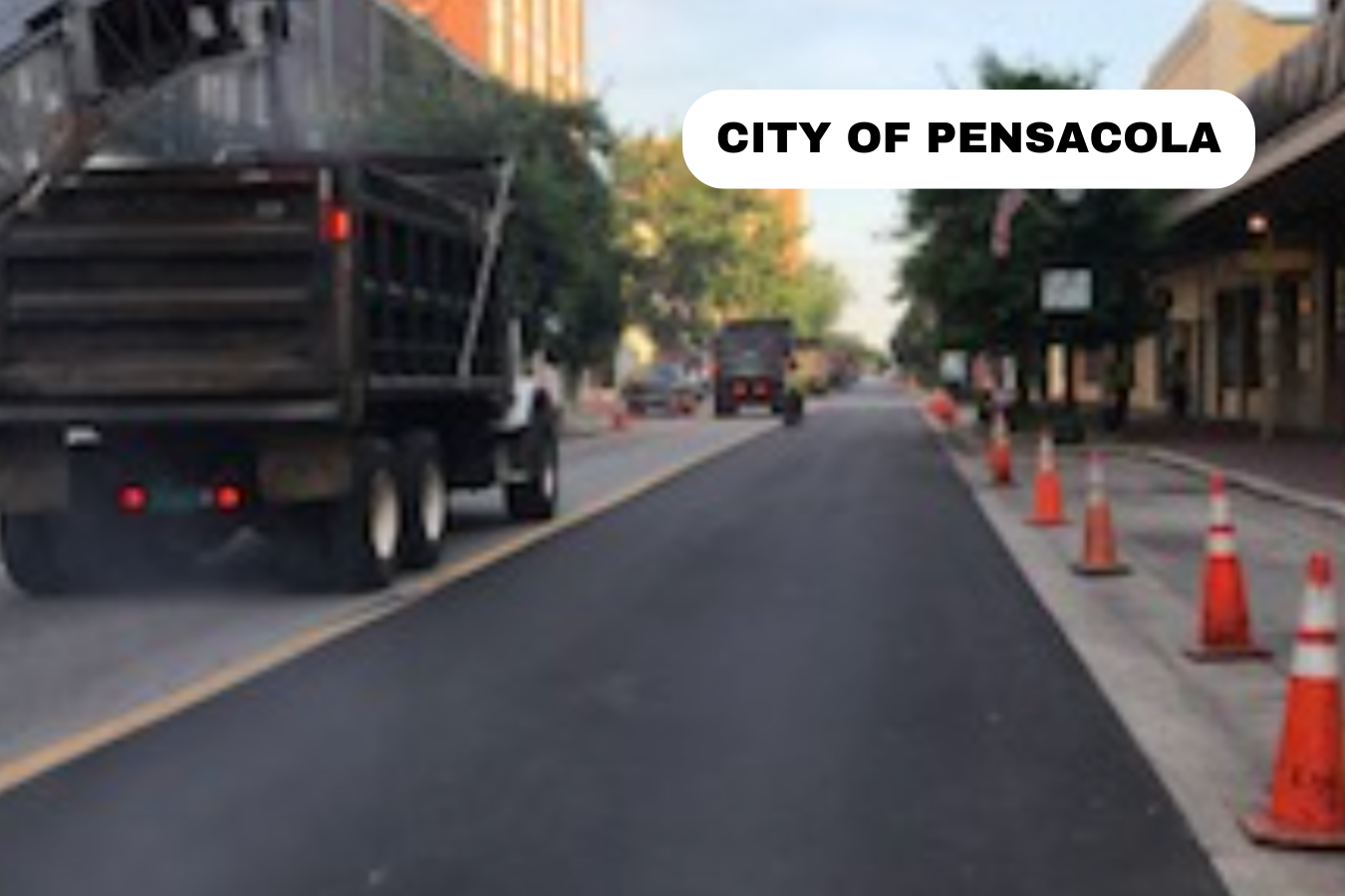 A street scene in Pensacola with construction cones on the sidewalk, trucks, and buildings lining the street.