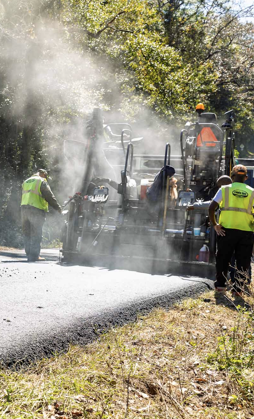 Workers in safety vests operating a road paving machine with smoke and dust, surrounded by trees and a sunny sky.