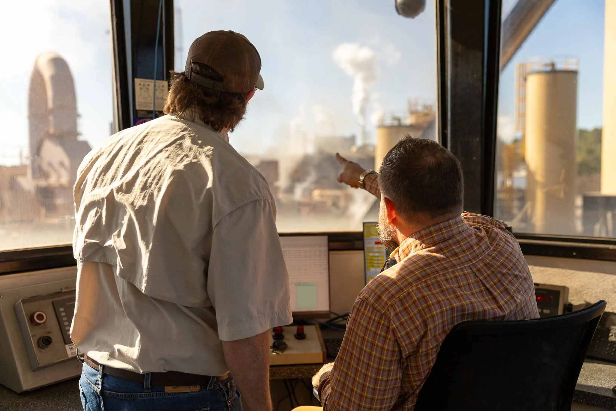 Two workers in an industrial control room, one pointing towards the outside view with factories and steam emitting from chimneys.