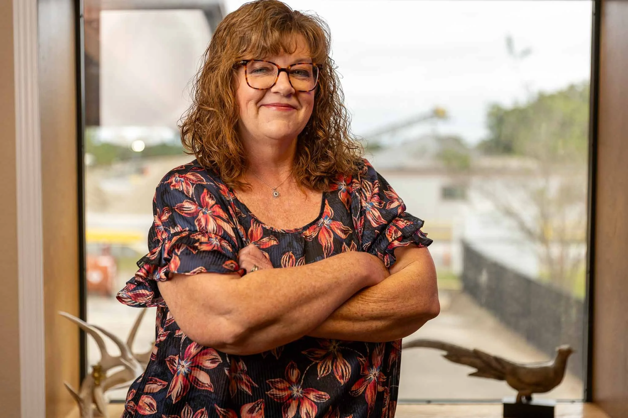 A woman with glasses, shoulder-length wavy hair, wearing a floral top, standing with arms crossed inside a building near a window, smiling at the camera.
