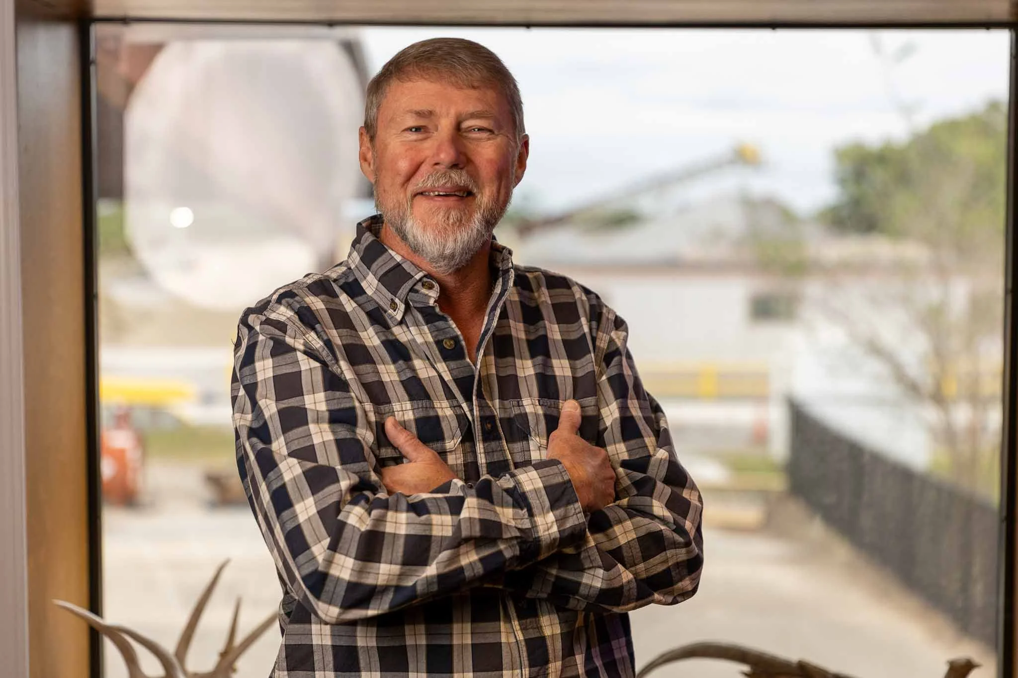 A smiling middle-aged man with gray hair and a beard, wearing a brown and black plaid shirt, stands with crossed arms in front of a window with an outdoor scene in the background.