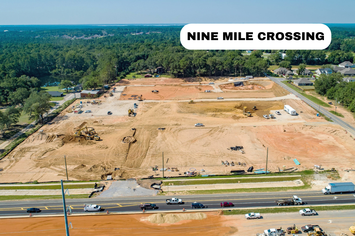 Aerial view of a construction site for a new development with excavators, construction vehicles, and dirt. Surrounding the site are residential neighborhoods and a busy road with traffic.