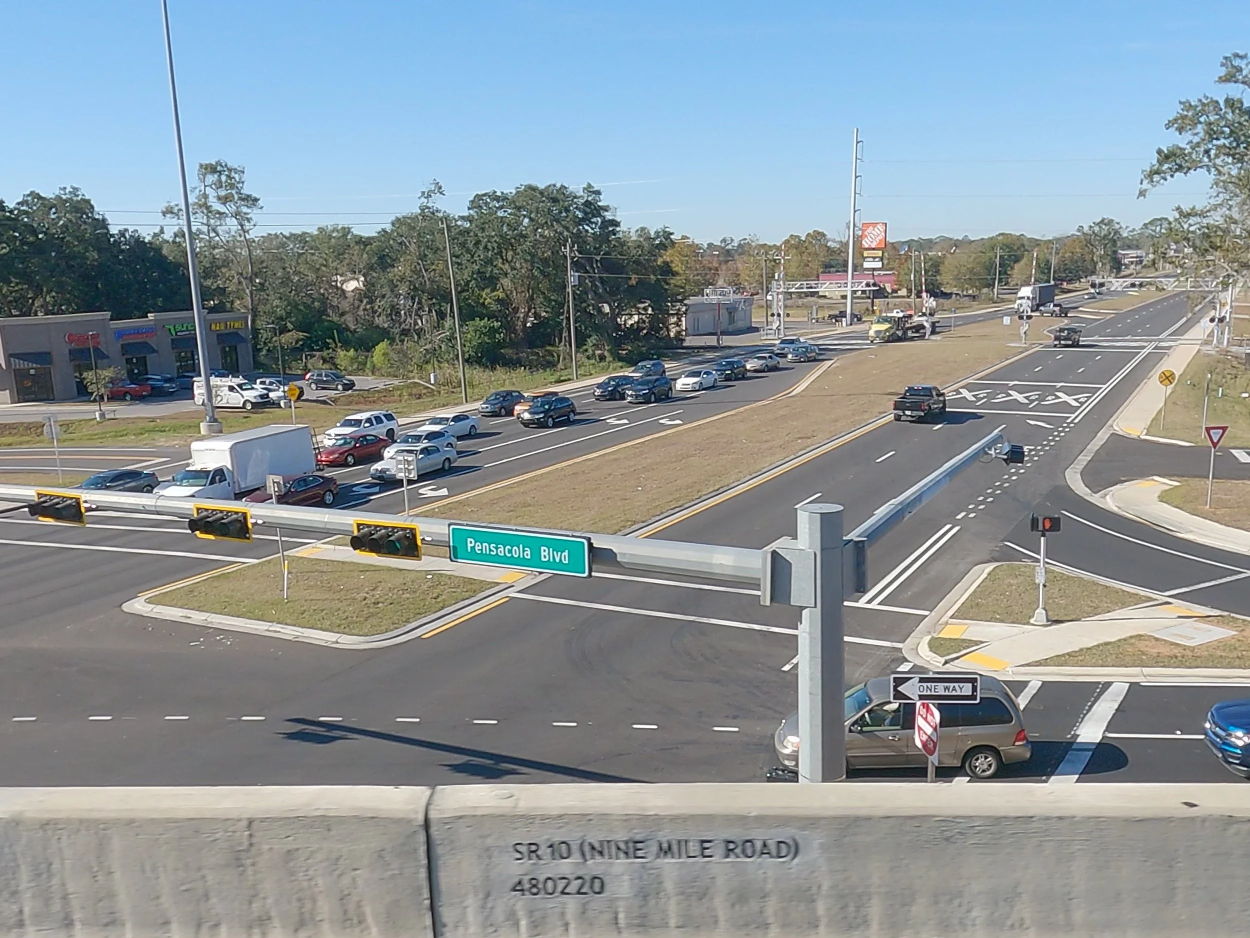 A busy intersection with multiple traffic lanes and cars, with a green street sign reading 'Pensacola Blvd' and a concrete barrier in the foreground. There are traffic lights and a directional sign indicating a one-way street, with some commercial bu