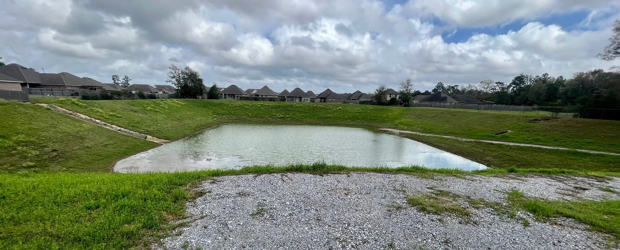 A small pond surrounded by grassy banks in a suburban neighborhood with houses and a fence in the background under cloudy skies.