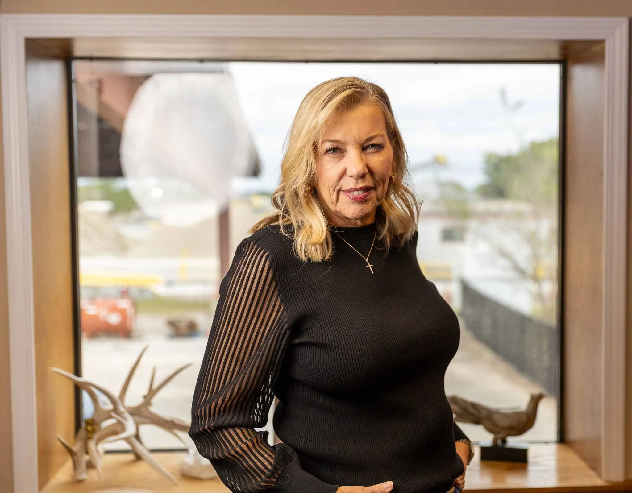 A middle-aged woman with blonde hair wearing a black top with striped, semi-transparent sleeves, standing indoors in front of a window with a view of the outdoors. There are decorative antlers and a wooden bird sculpture on a table behind her.
