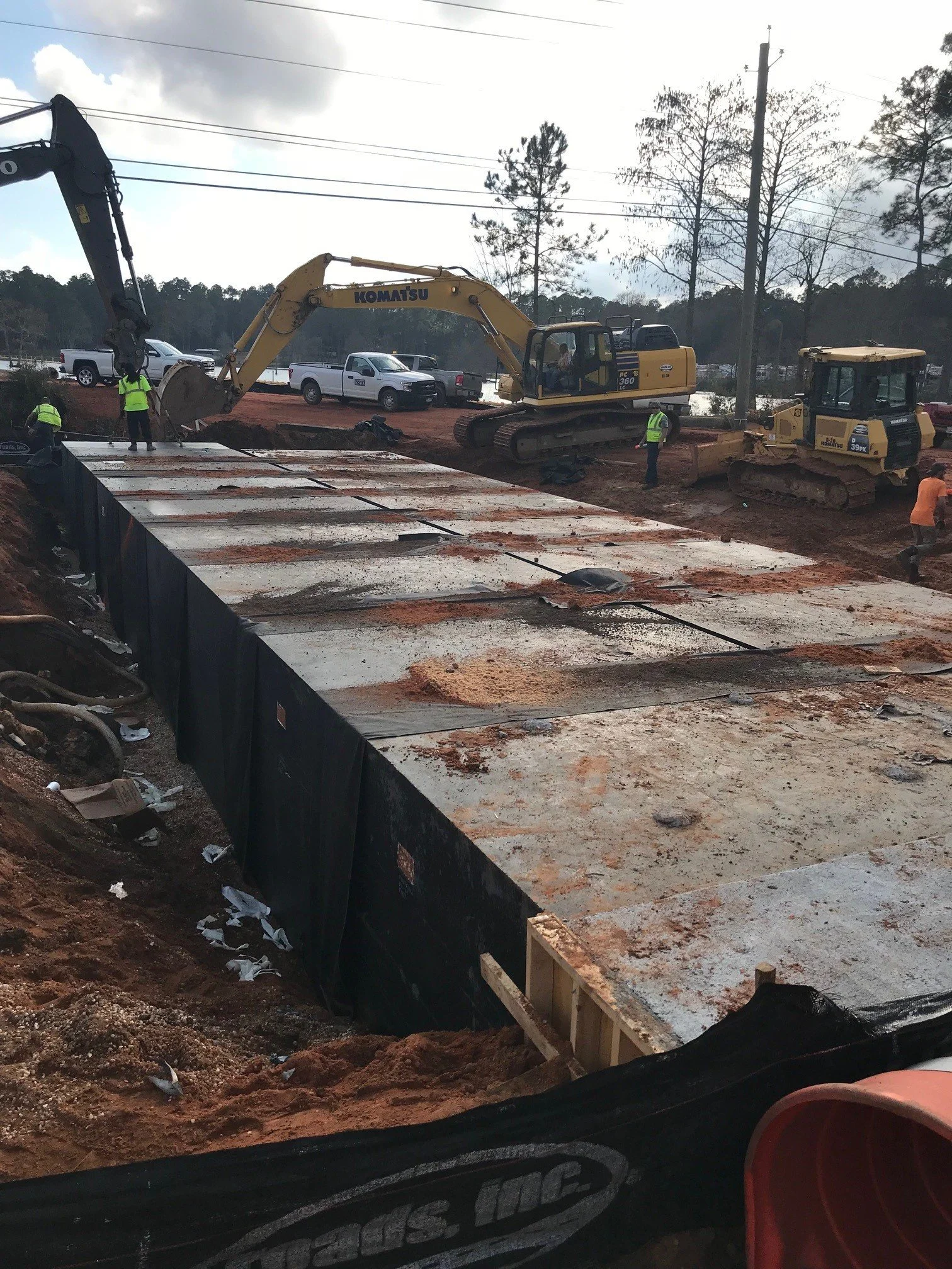 Construction site with excavators and workers installing a large concrete slab on a dirt base, with vehicles and trees in the background.