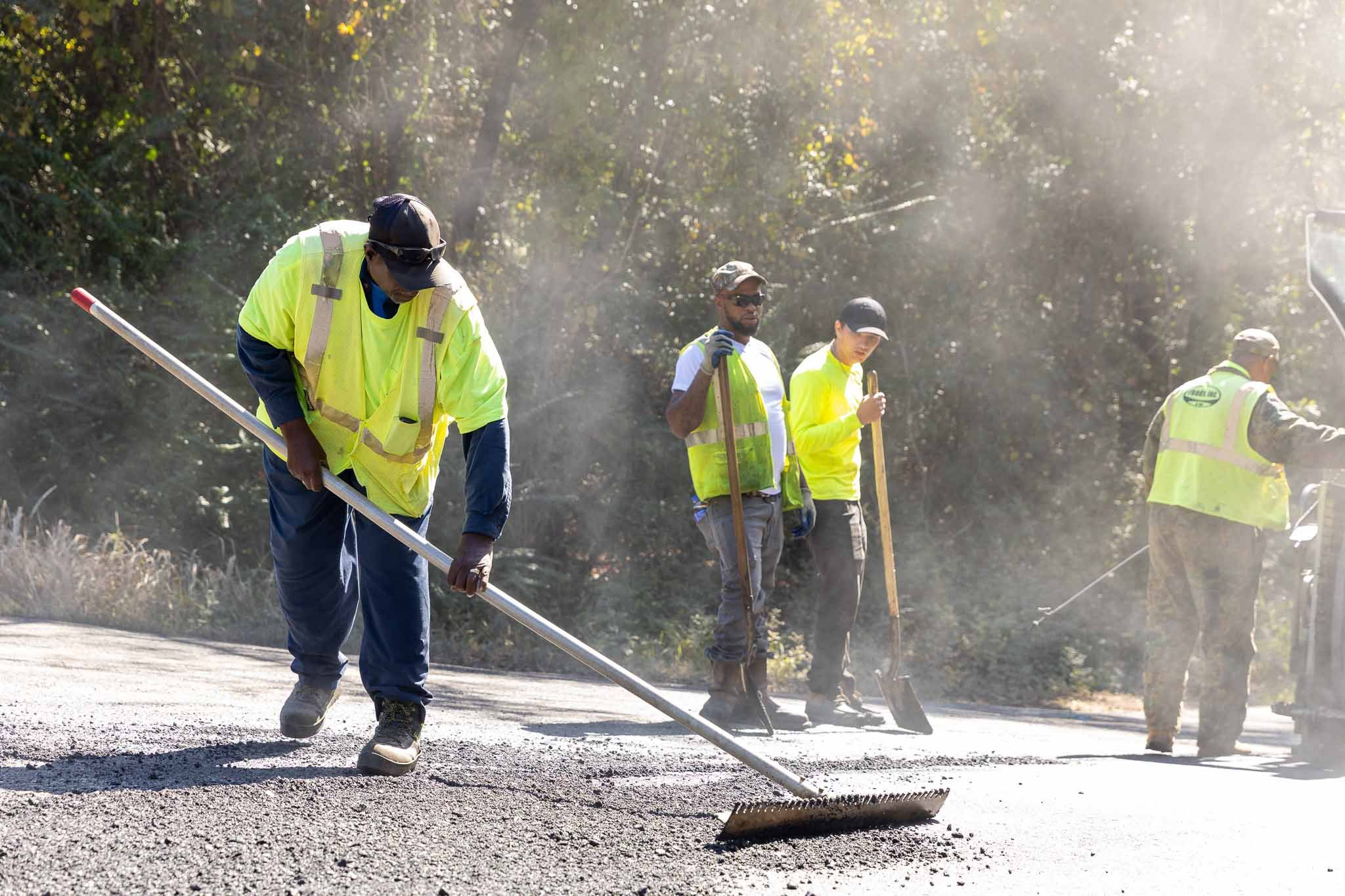 Construction workers in reflective vests working on a road repair project amidst dust and greenery.