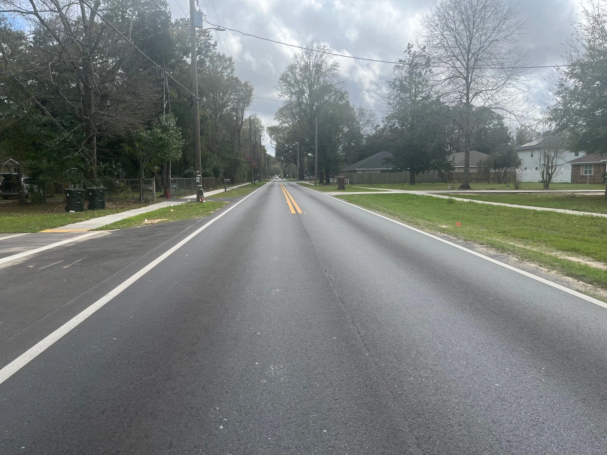 Empty residential street with double yellow lines, sidewalks on both sides, and trees lining the street. Overcast sky.