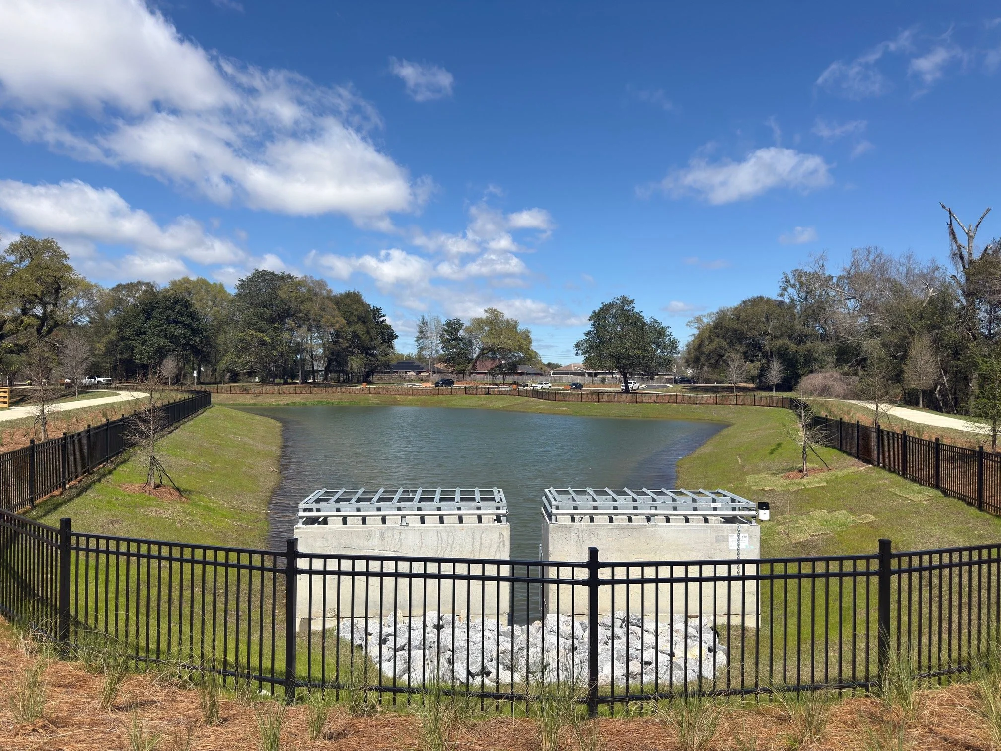 Reservoir with two concrete outlet structures, surrounded by a black metal fence and trees, under a partly cloudy blue sky.
