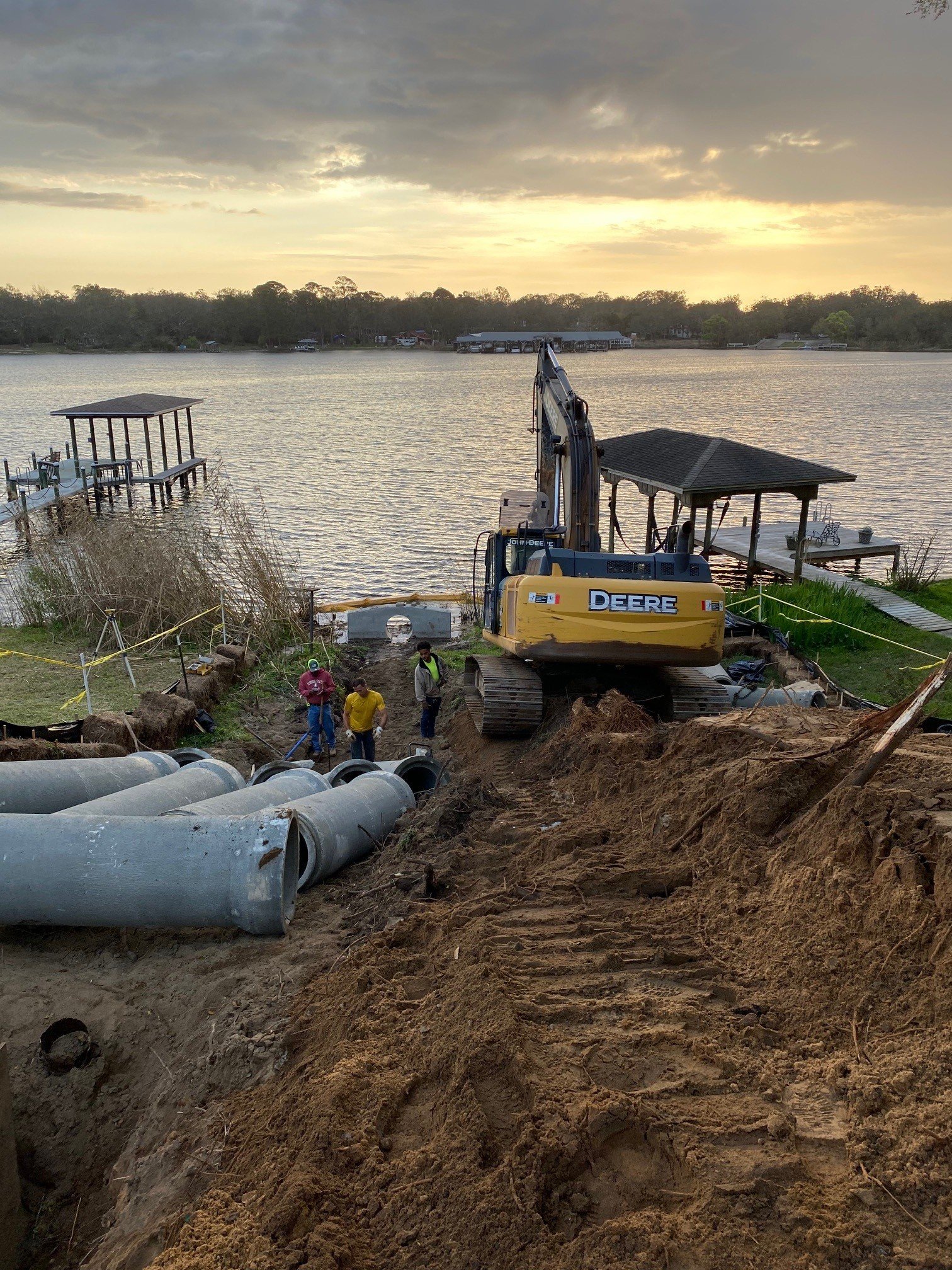 Construction workers and a yellow Deere excavator working on installing large concrete pipes near a lakeside or river, with a gazebo and docks in the background at sunset.