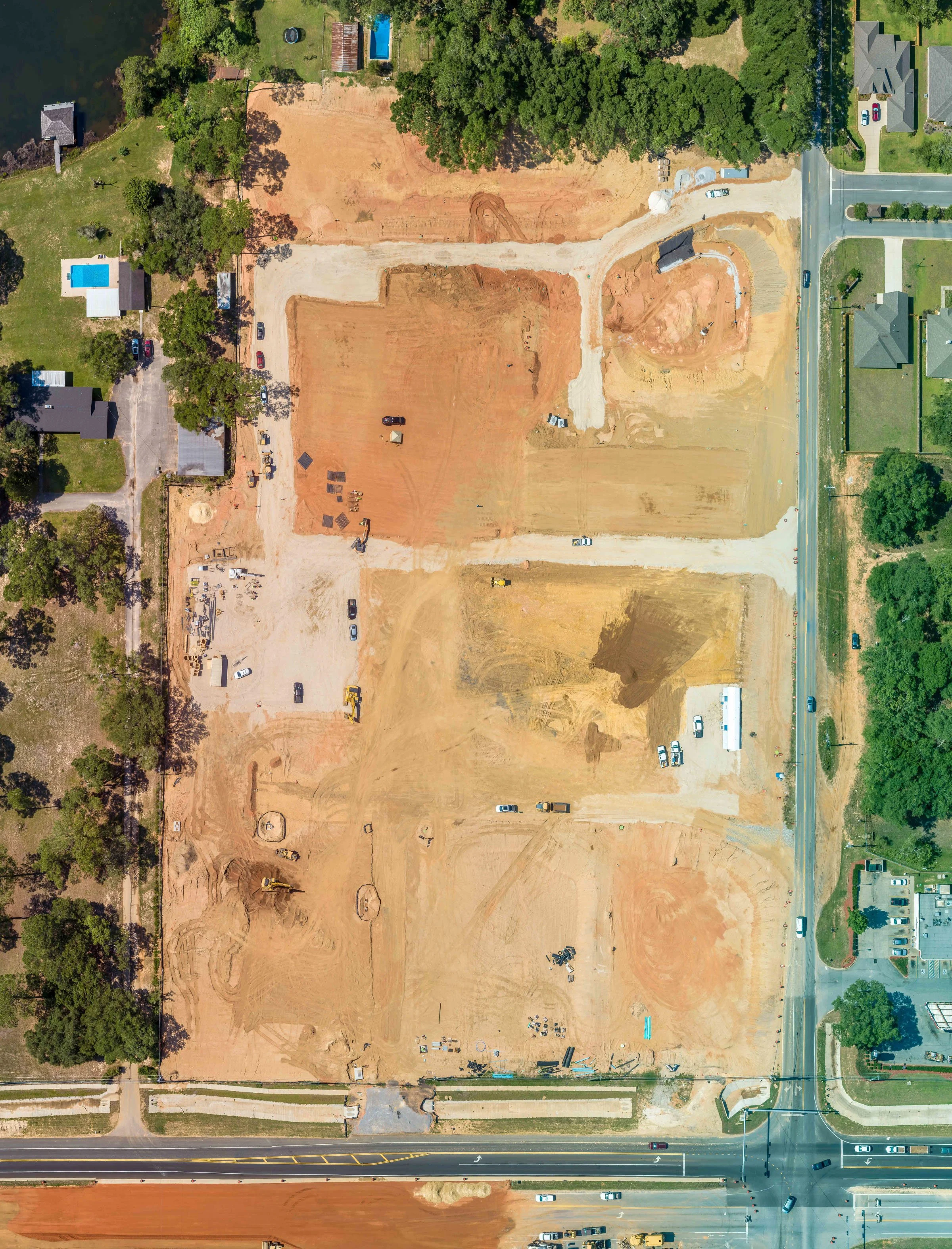 Aerial view of a construction site with cleared earth, construction vehicles, and equipment, surrounded by trees and neighboring houses, with roads at the bottom and right edges.