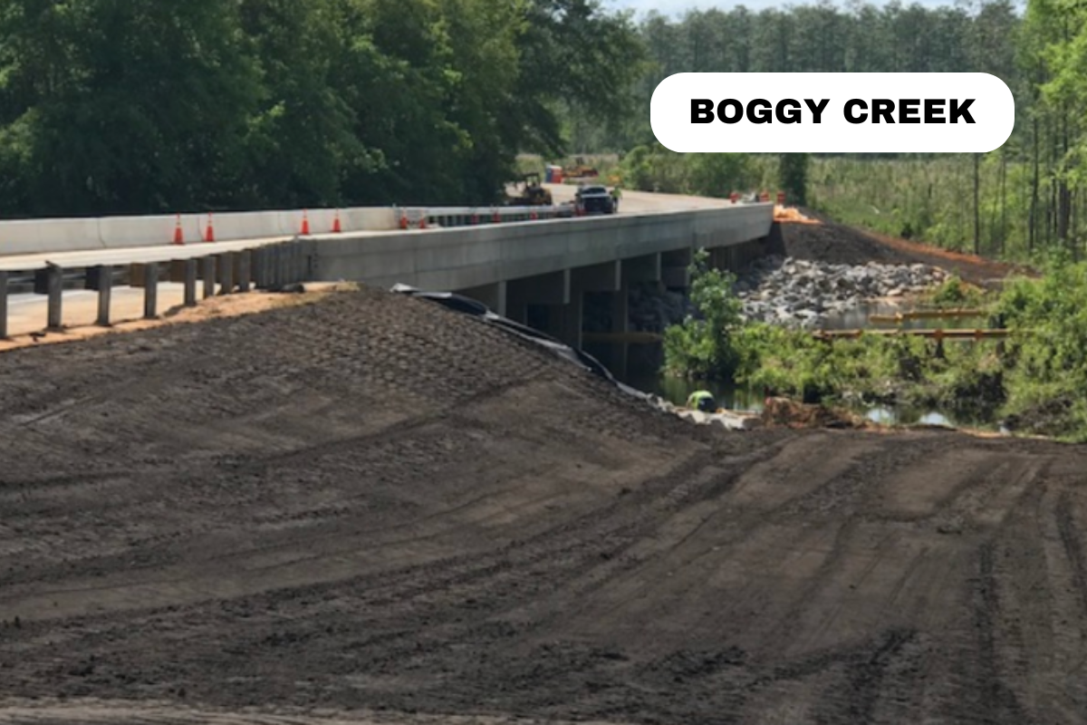 Construction site on a bridge over Boggy Creek, with construction vehicles and orange cones, dirt road in foreground, trees in background.