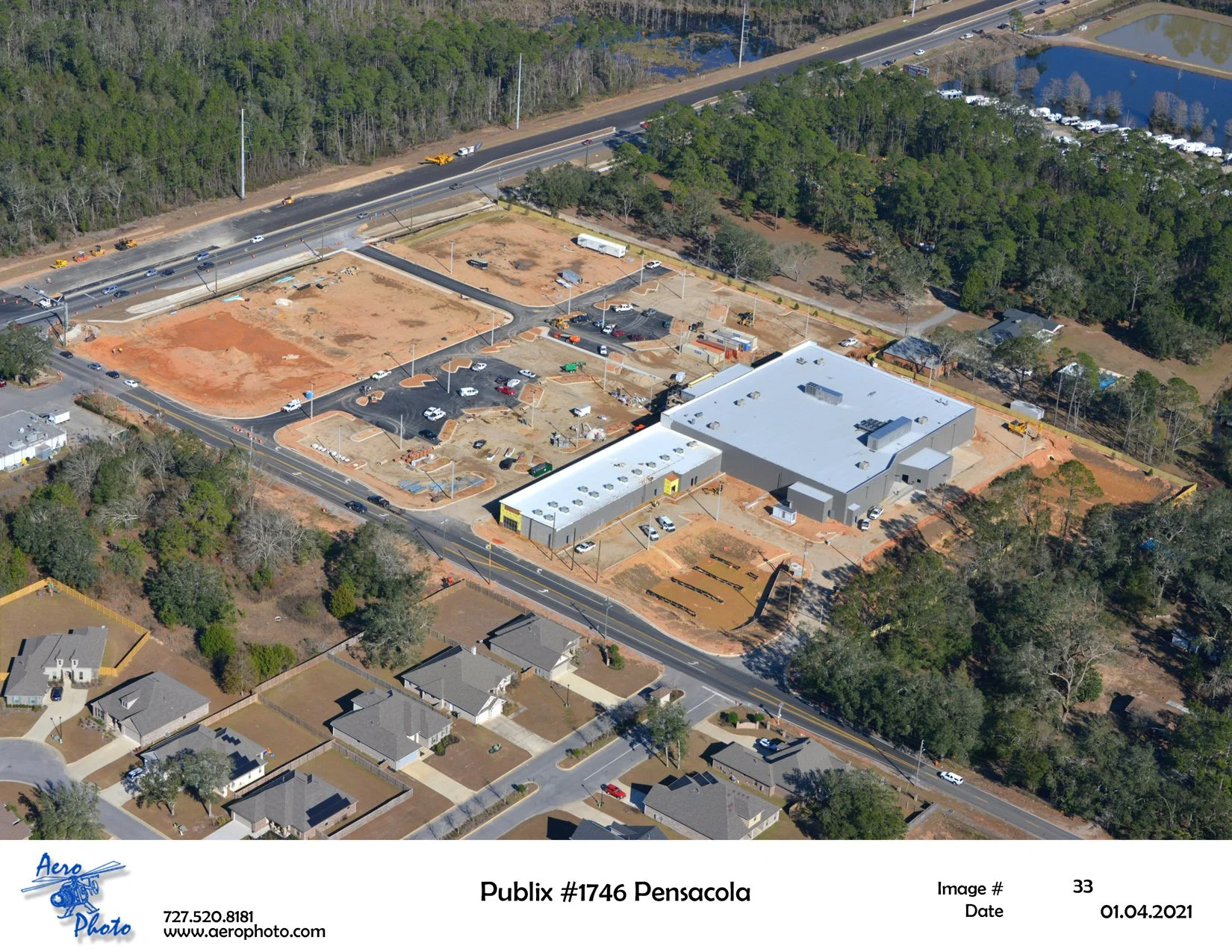 Aerial view of a parking lot and a grocery store under construction next to a highway, with residential houses nearby and wooded areas in the background.