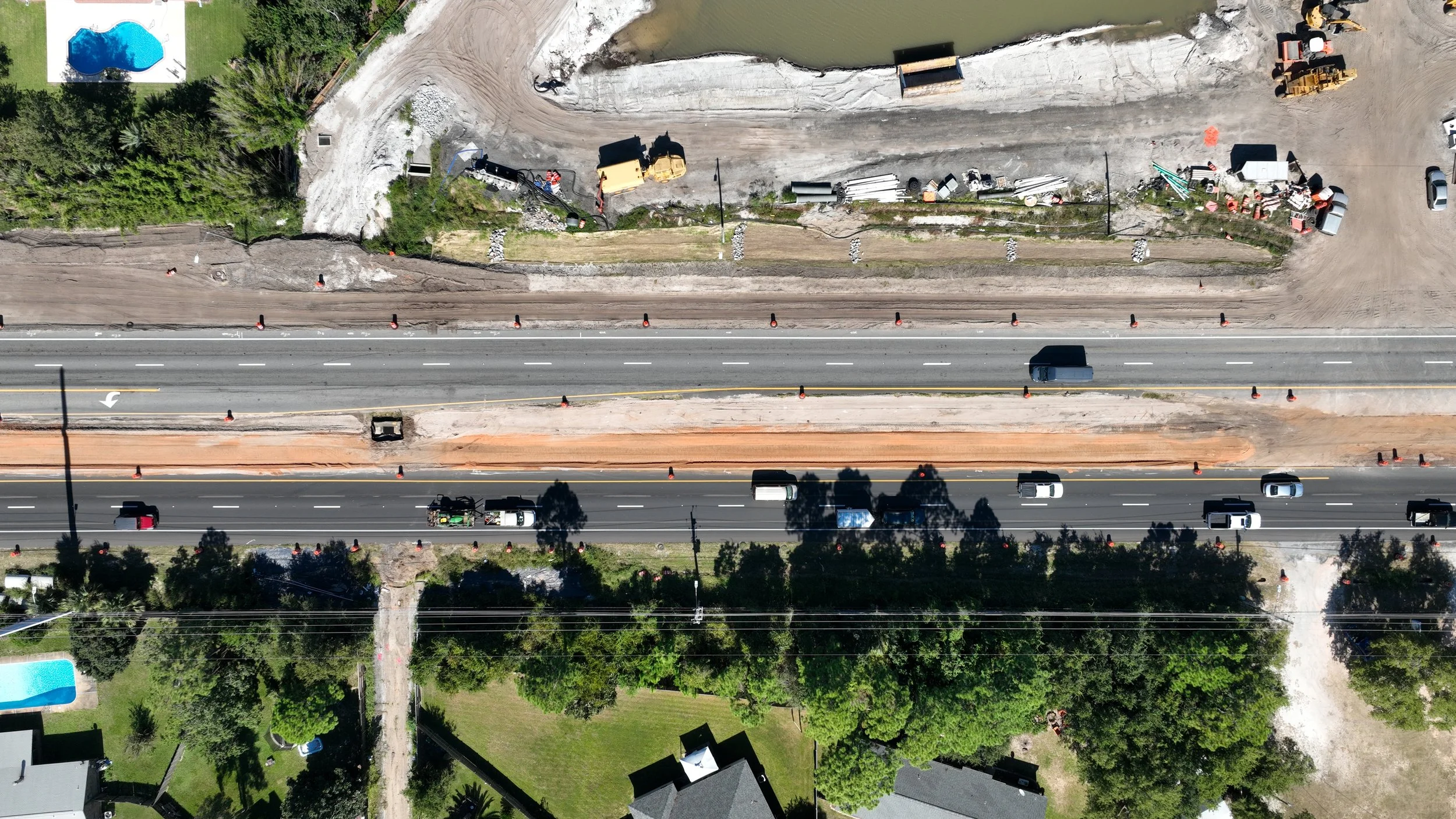Aerial view of a multi-lane road under construction, with lanes partially paved, construction vehicles, and barriers, surrounded by greenery and residential houses.