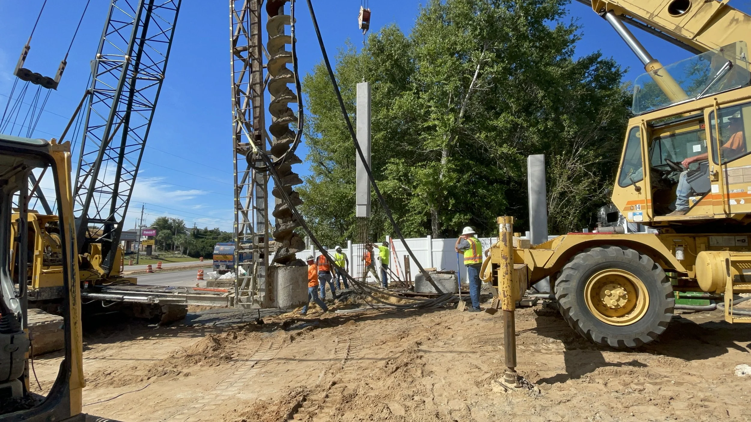 Construction workers and heavy machinery working at a site, with a bright blue sky, trees, and a white fence in the background.