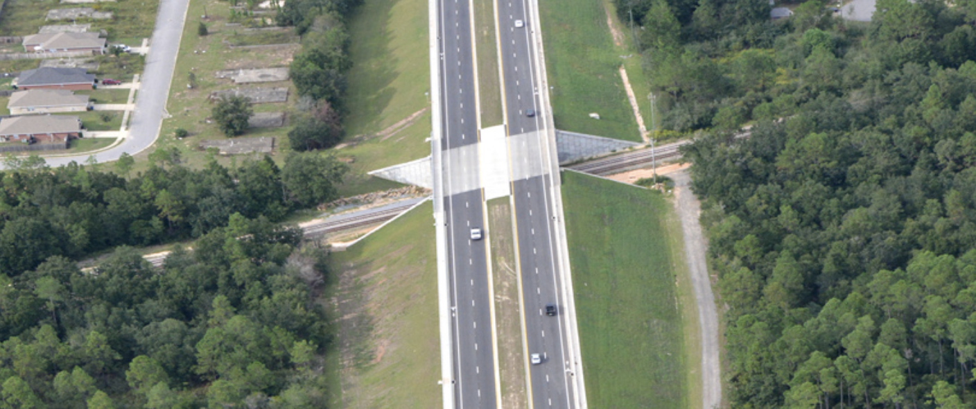 Aerial view of a highway overpass crossing through a green forest with a residential neighborhood nearby.