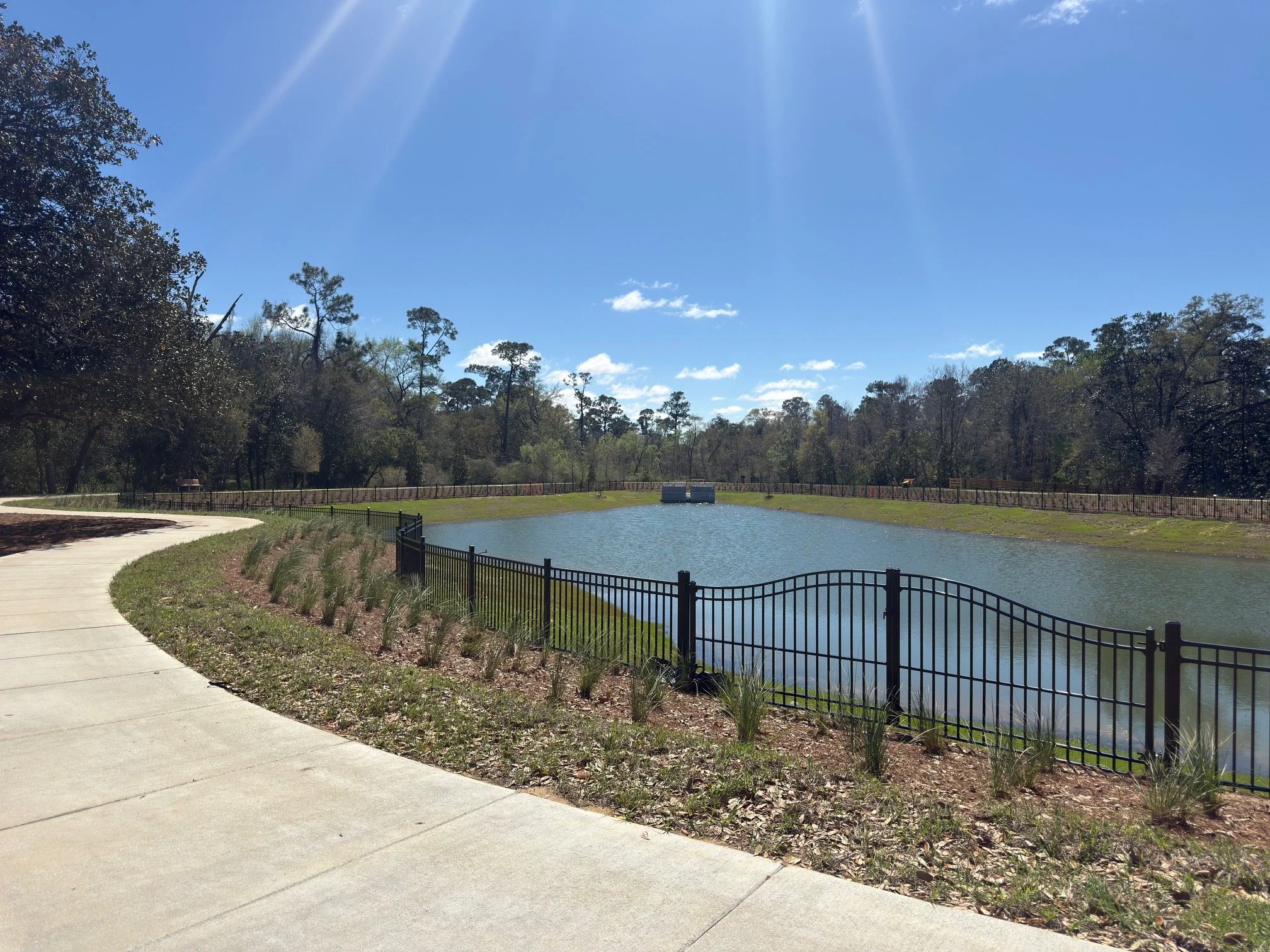 A park pathway curves alongside a small pond, surrounded by a black metal fence. Trees and blue sky with clouds are visible in the background, and the sun shines brightly overhead.