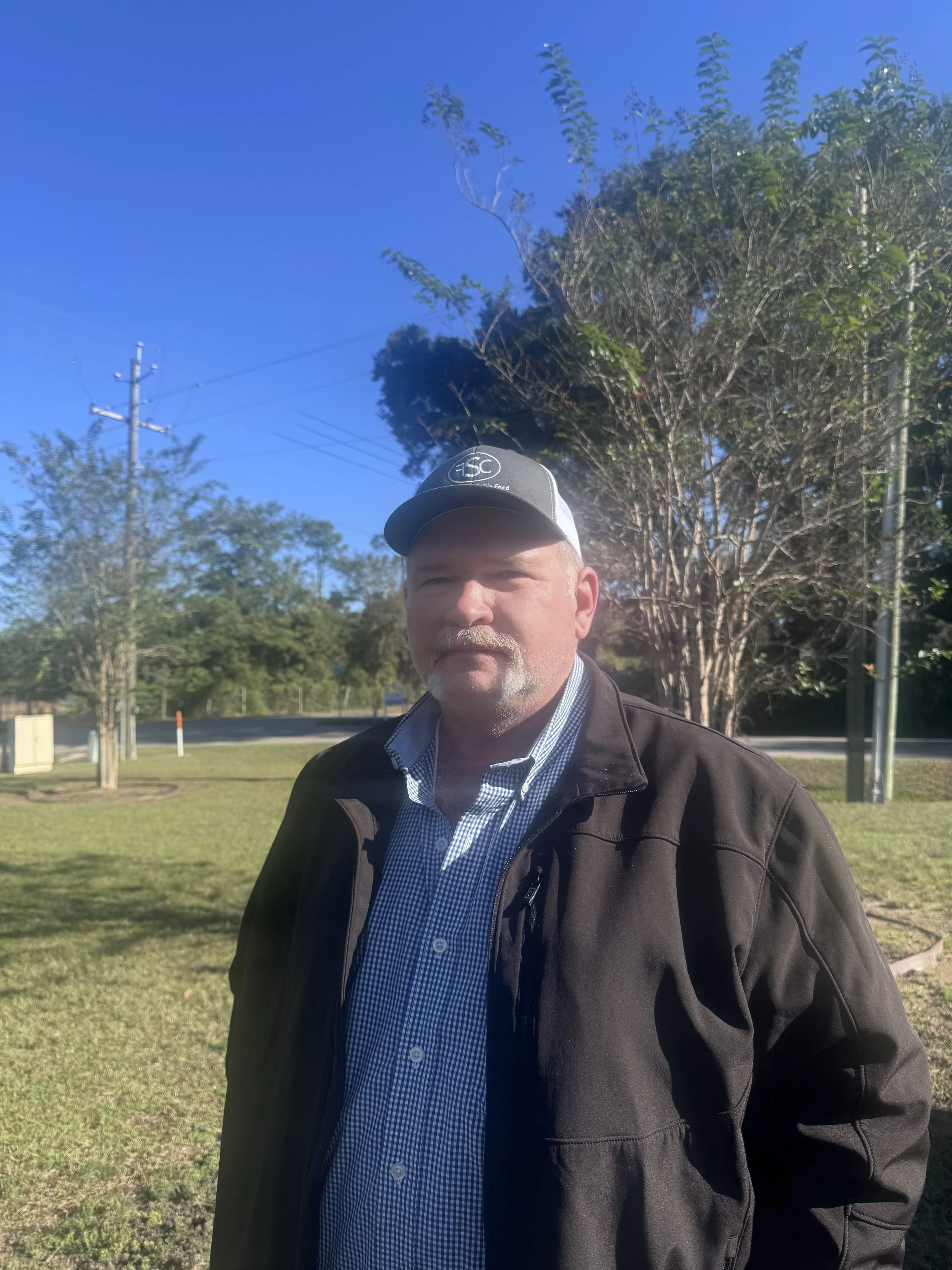 A man standing outdoors in front of a tree, wearing a black jacket, checkered shirt, and a black and white baseball cap.